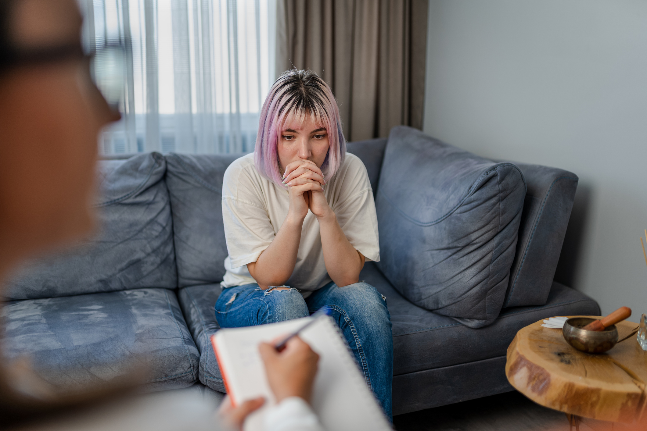 Woman listens intently on sofa in therapy session, therapist in foreground with notepad