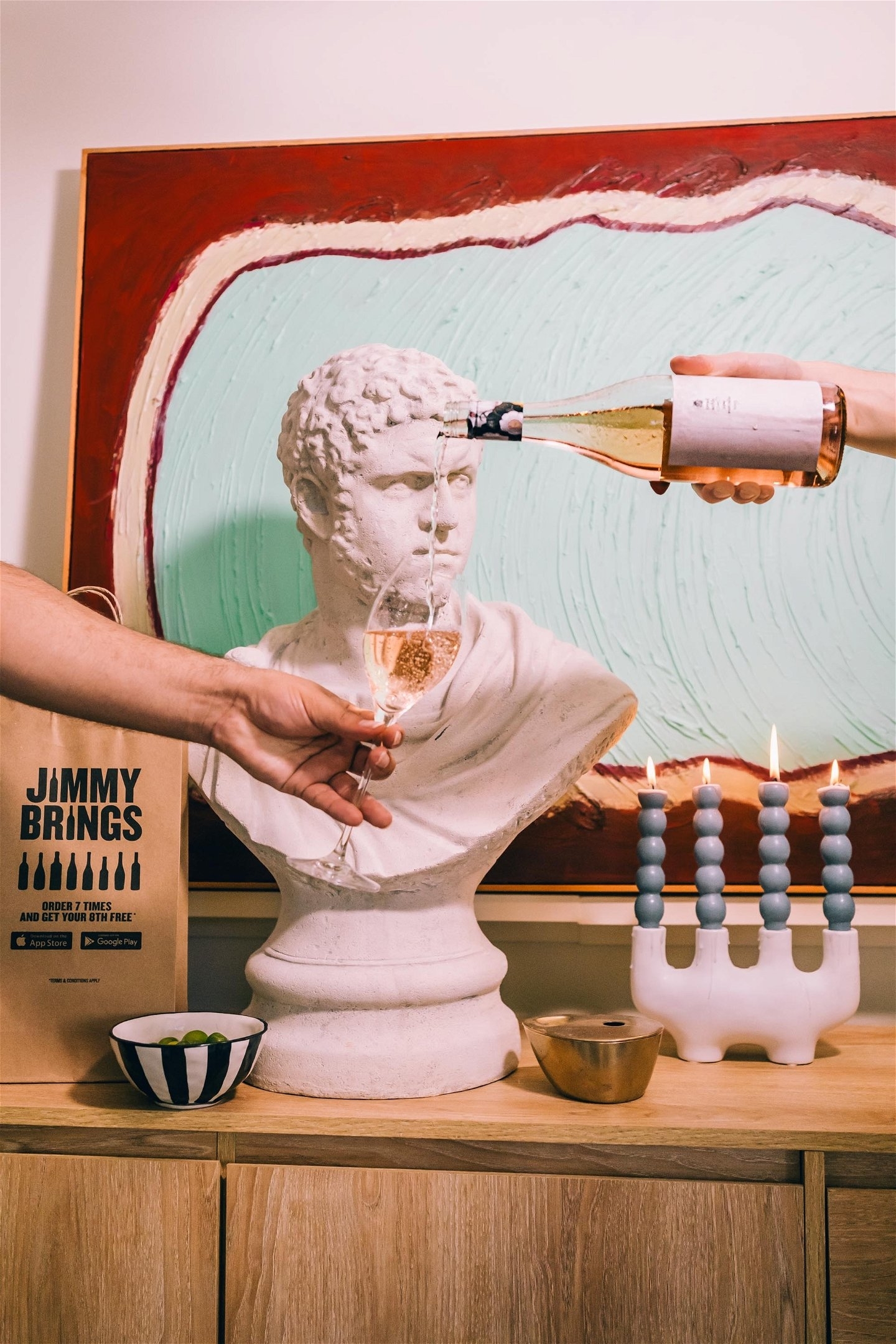 Person pours sparkling wine from a bottle into a glass held by a bust sculpture, with candles and a book nearby