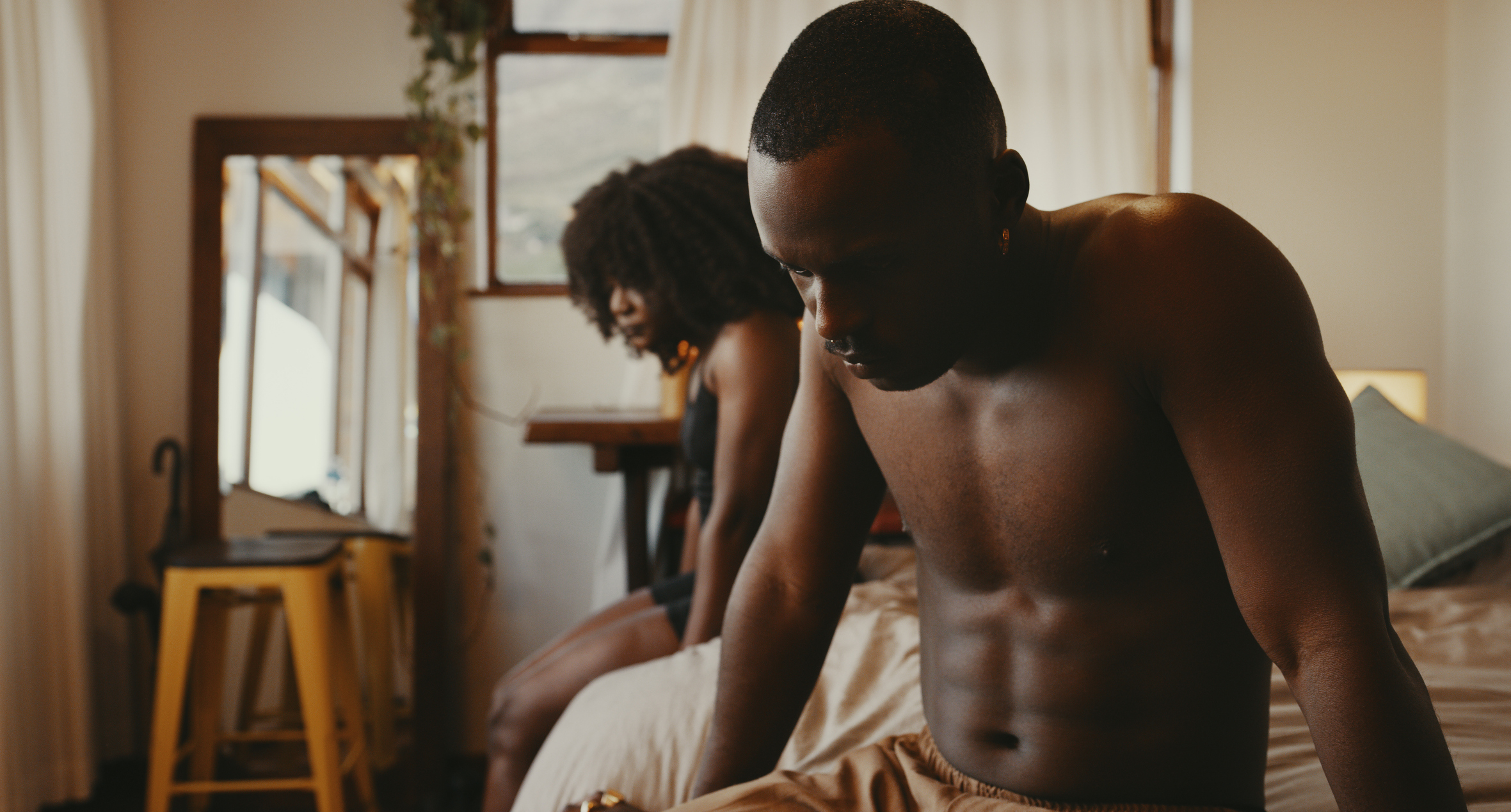 A couple in a contemplative moment, with a woman seated at the bed&#x27;s edge and a man standing turned away