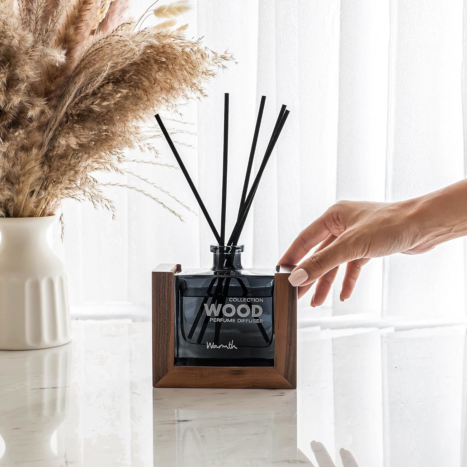 A person&#x27;s hand adjusting reed diffusers in a glass bottle with a wooden base on a sunlit window sill