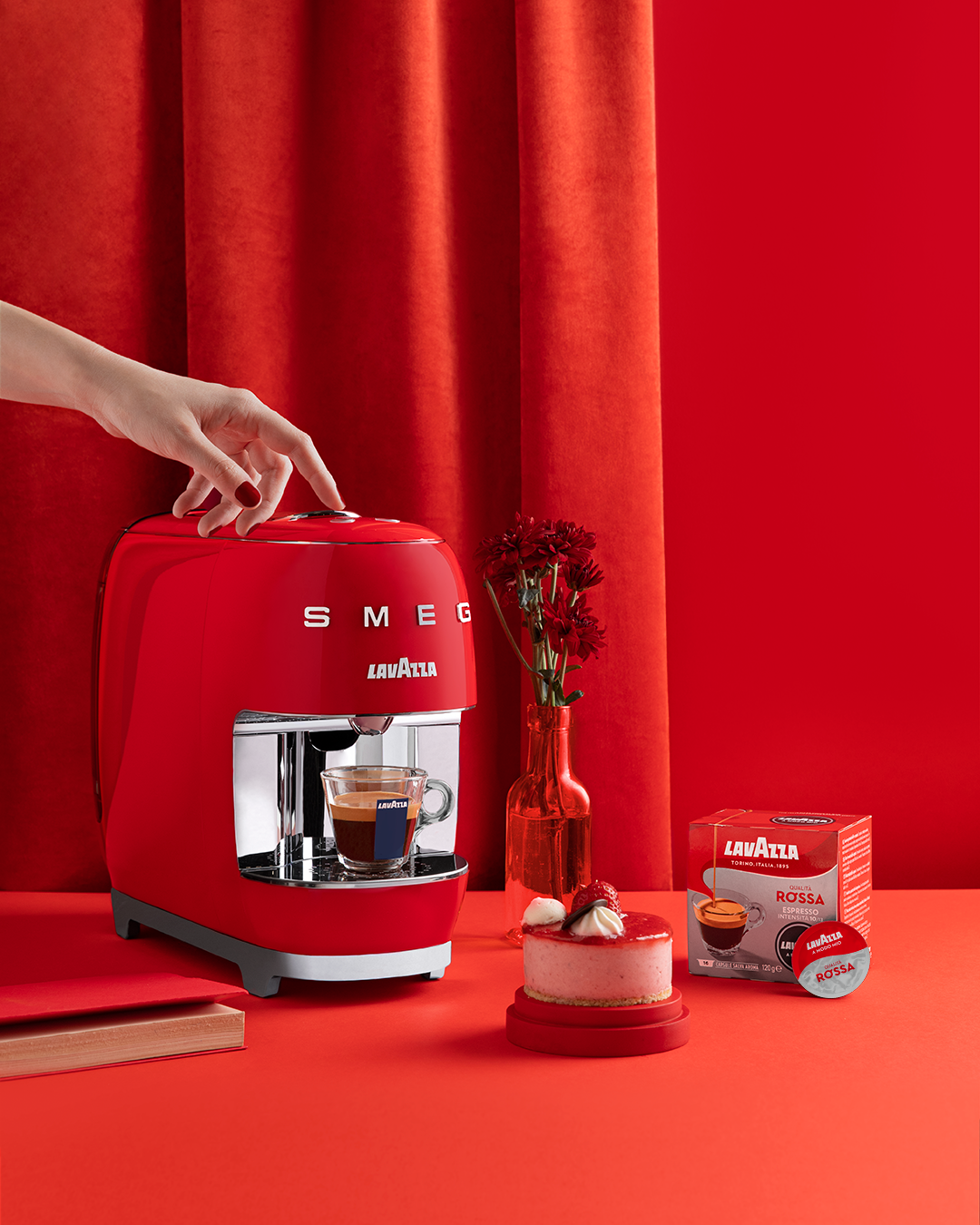 Person using a red SMEG espresso machine beside a cup of coffee, pastry, coffee beans, and flowers on a counter