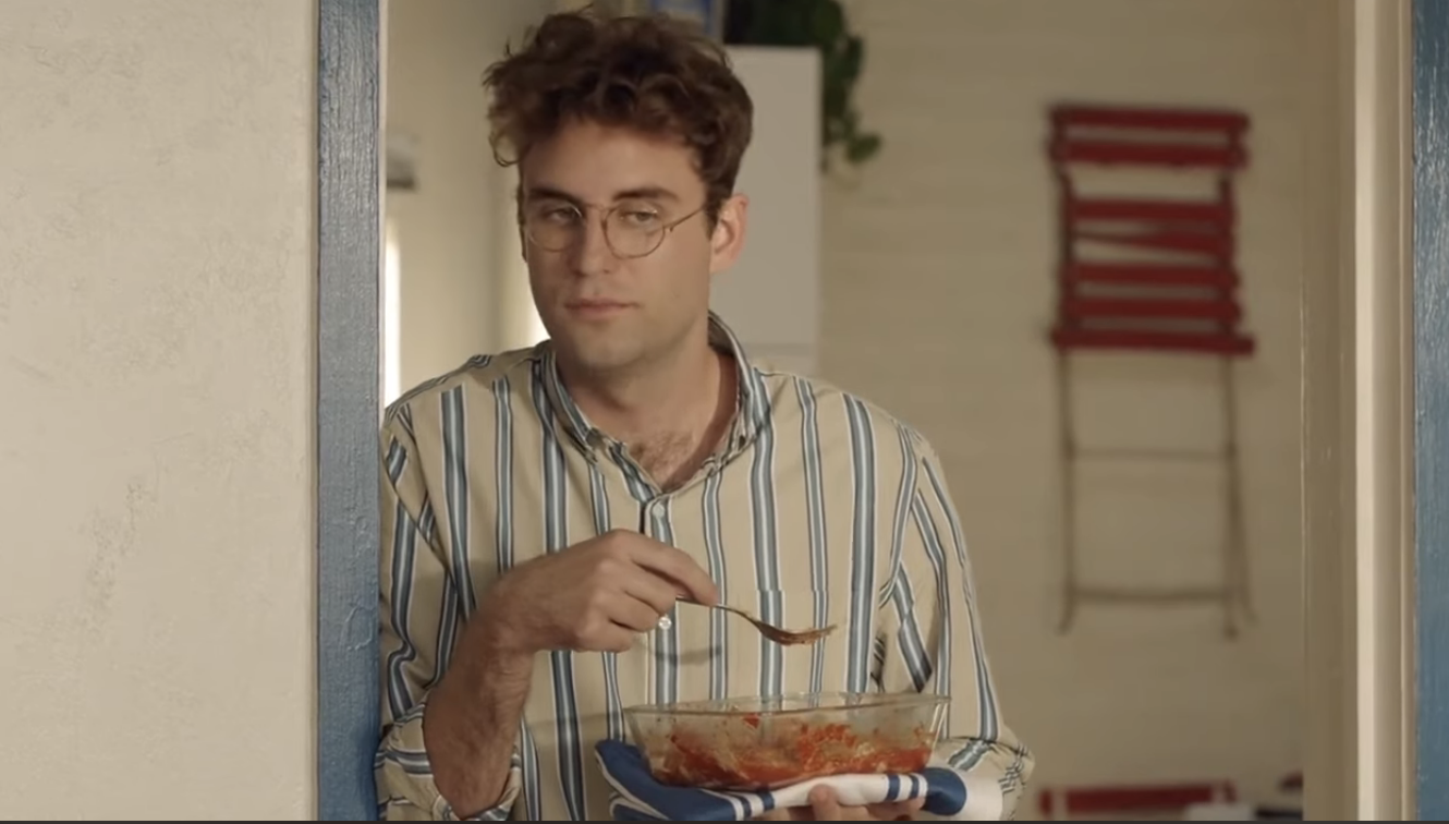 Man with curly hair wearing striped shirt eating from a bowl while standing in front of a mirror