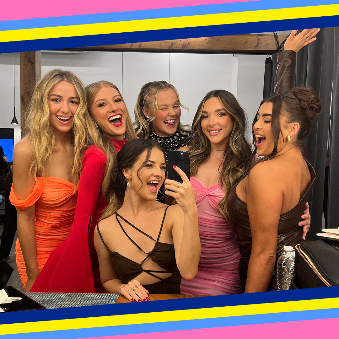 Group of people smiling for a selfie, some dressed in elegant gowns, one in a suit, in a room with a mirror