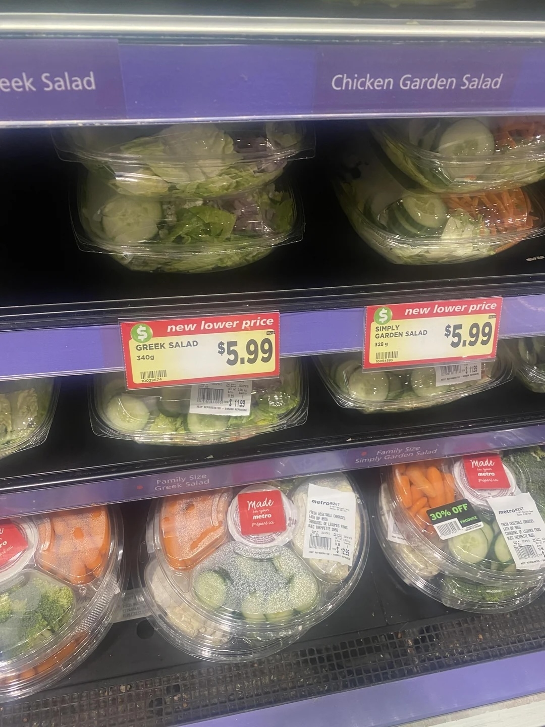 Supermarket shelf with various salads, new lower price labels at $5.99, Greek and Garden salads visible