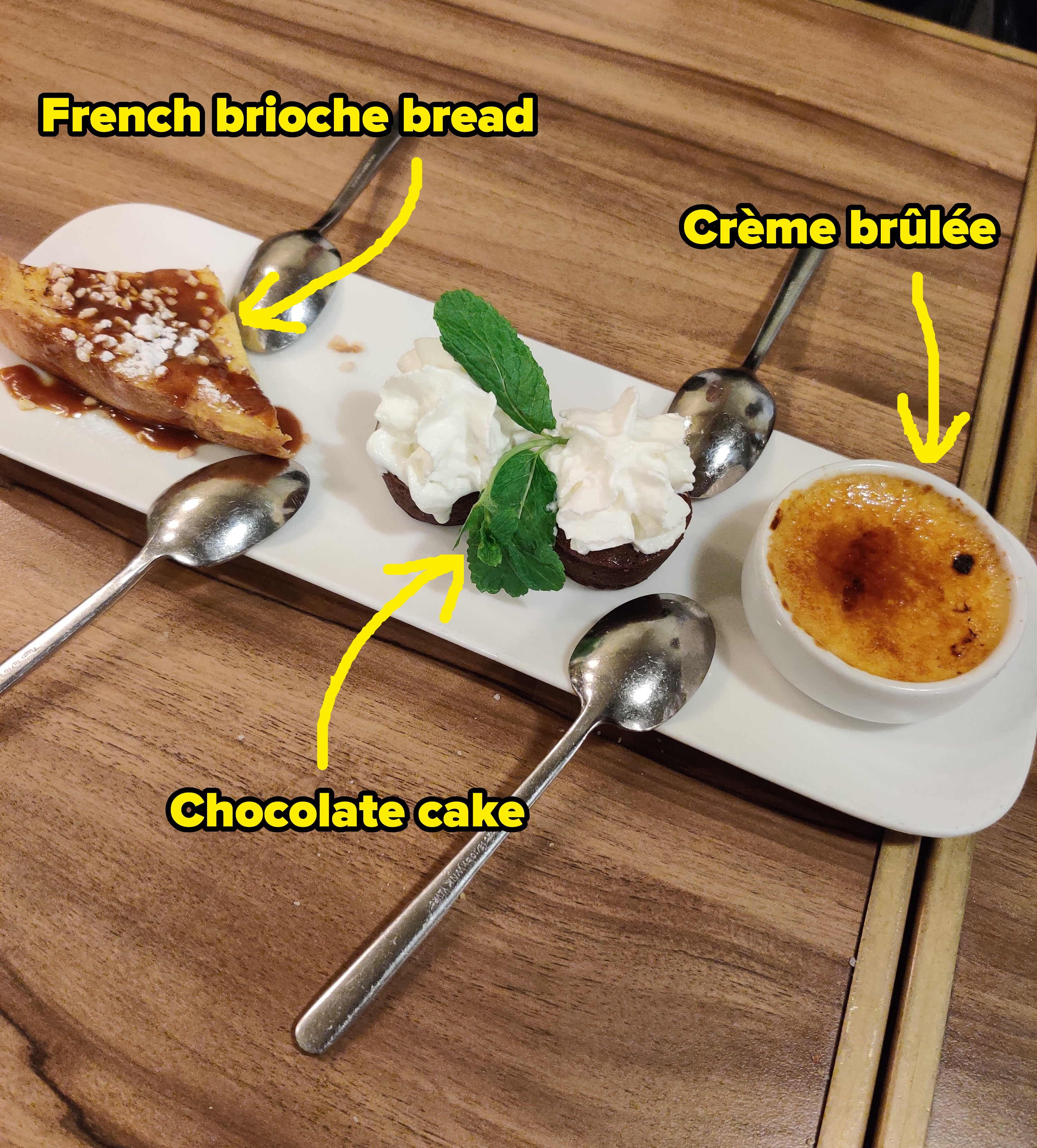 A tray with various desserts including a slice of cake, whipped cream, and a crème brûlée, with spoons, on a wooden table