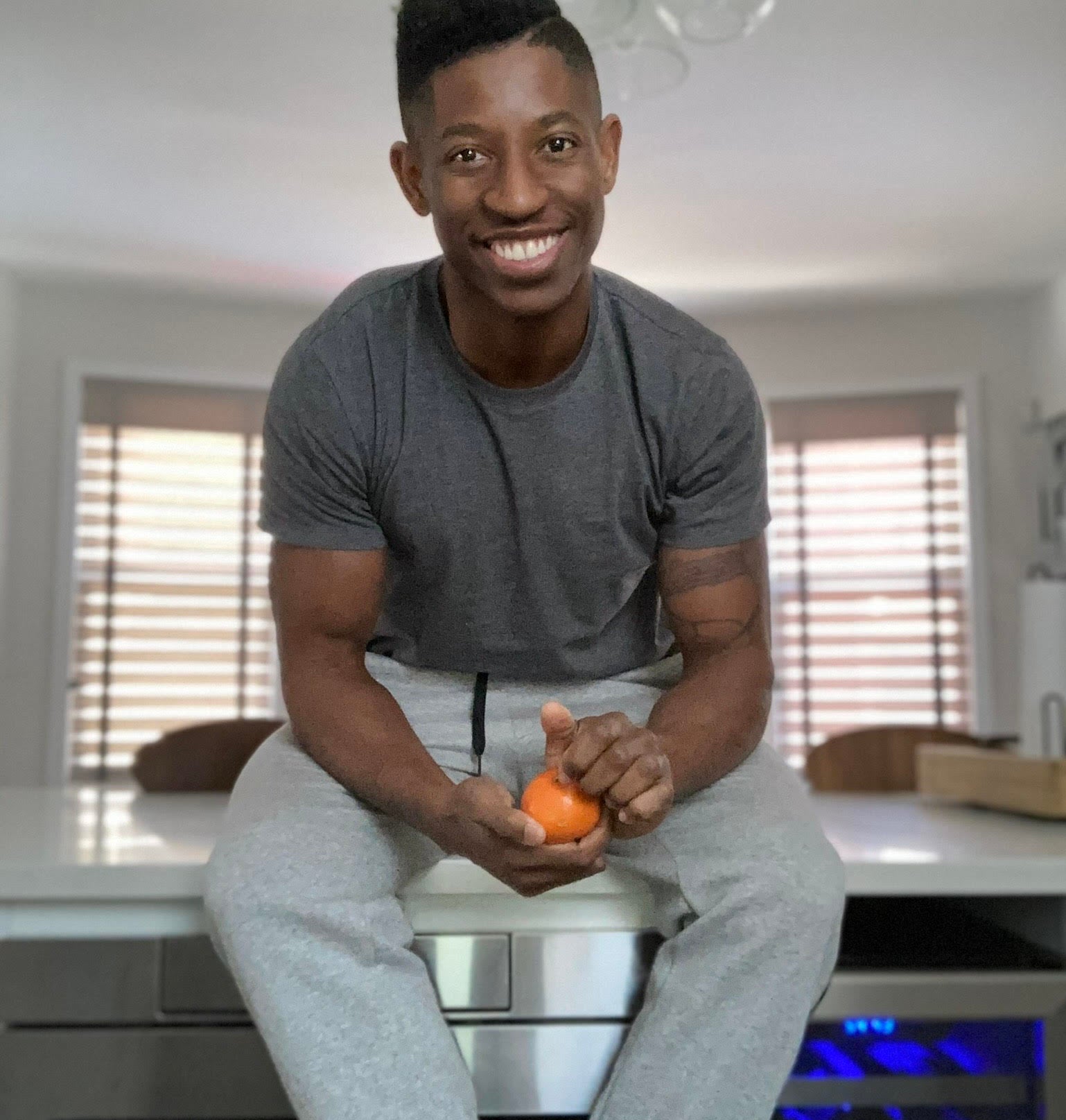 Man seated inside home, smiling at camera, holding an orange. He wears a T-shirt and jogging pants