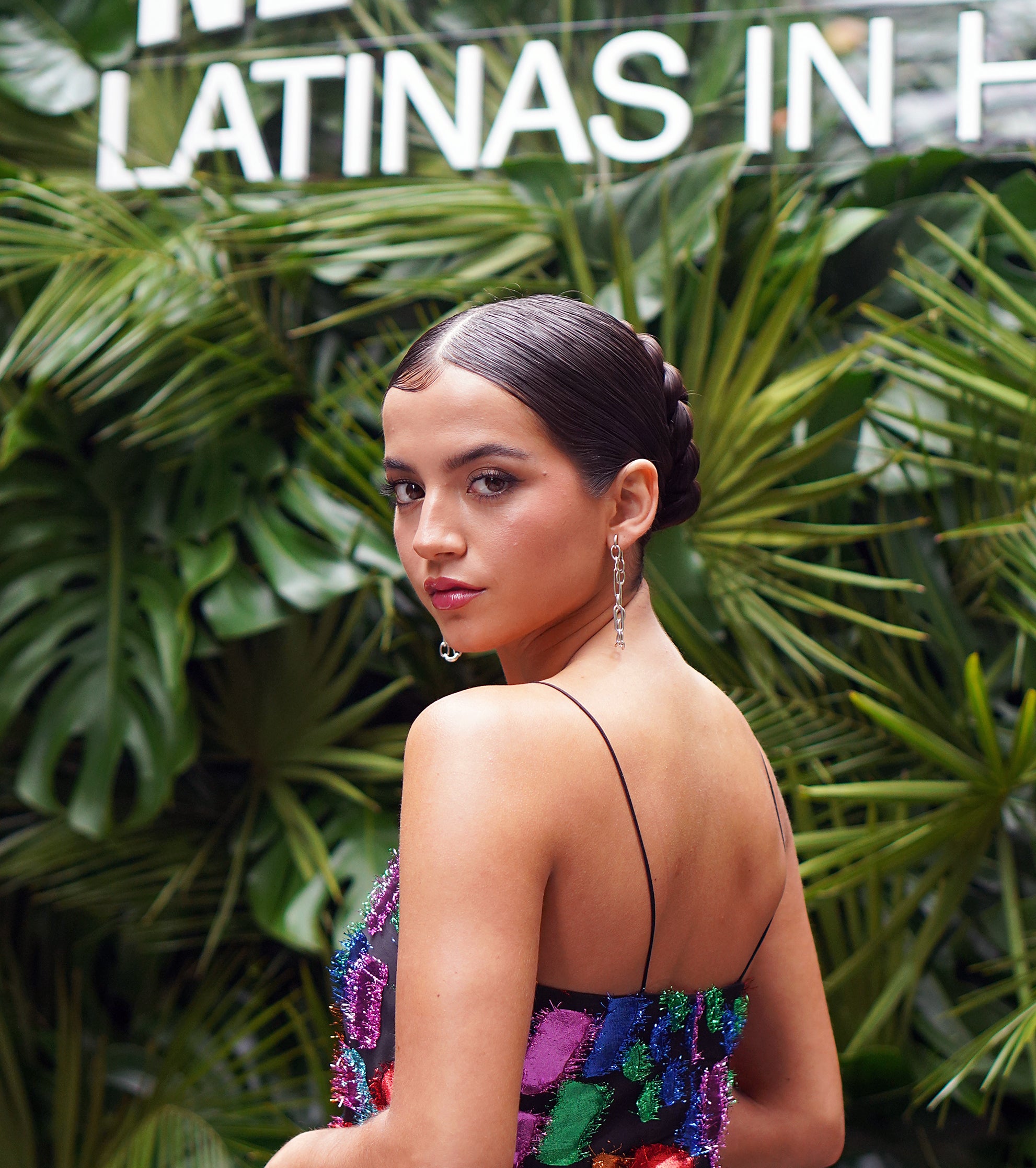 A woman poses in front of a backdrop with text, wearing a sequined outfit with a braid hairstyle