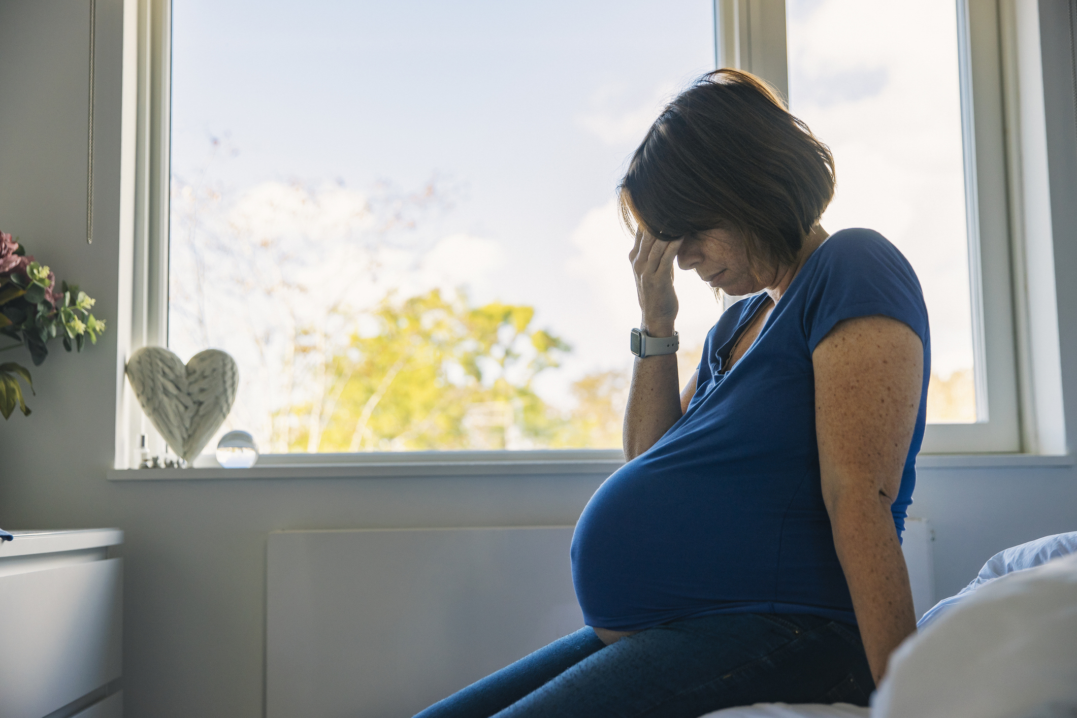 A pregnant woman in a blue shirt sits on a bed, looking down with her hand on her forehead, appearing stressed or deep in thought. A window is behind her