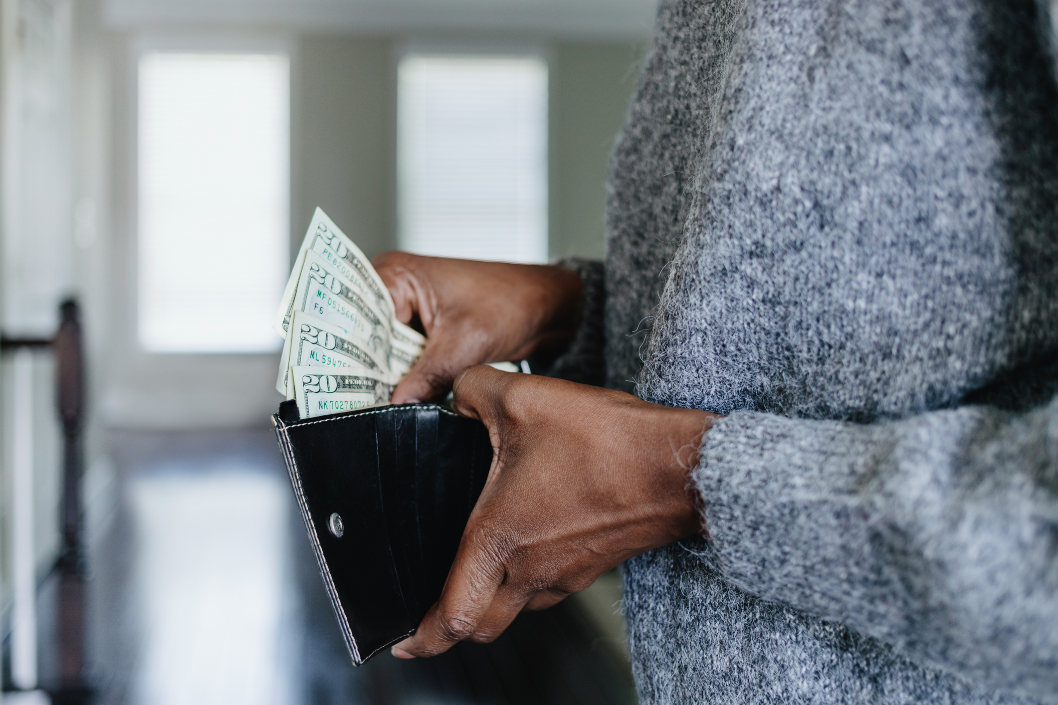 A person in a gray sweater is holding a black wallet and counting several U.S. dollar bills