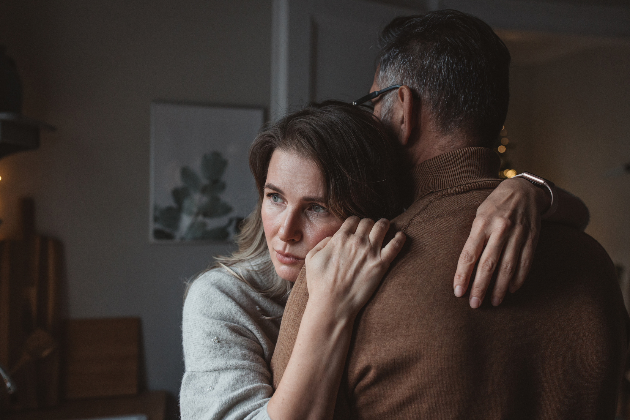 A woman hugs a man tightly, her expression serious and contemplative. They are indoors, suggesting a moment of support or comfort