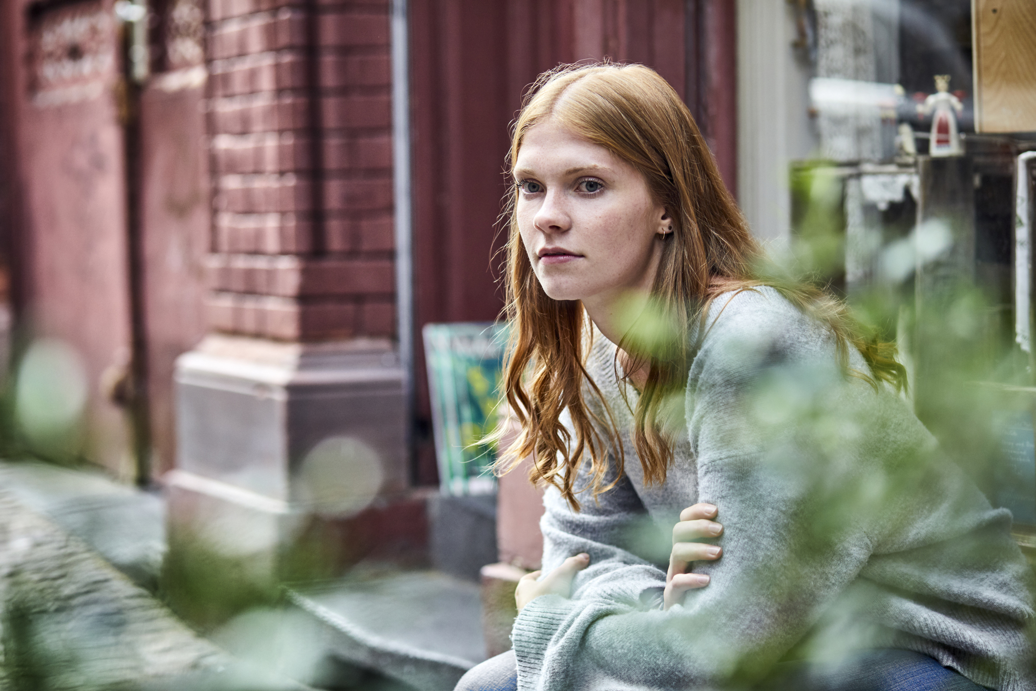 A woman with long red hair and a contemplative expression sits outside on a bench, wearing a casual sweater