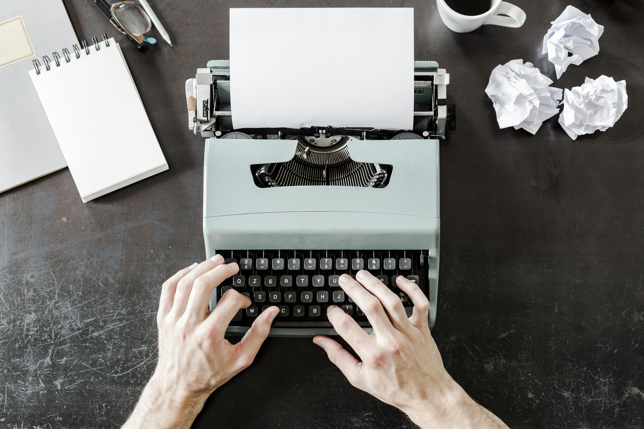 Hands typing on a vintage typewriter with a blank sheet of paper, surrounded by crumpled paper balls, a notebook, and a coffee cup