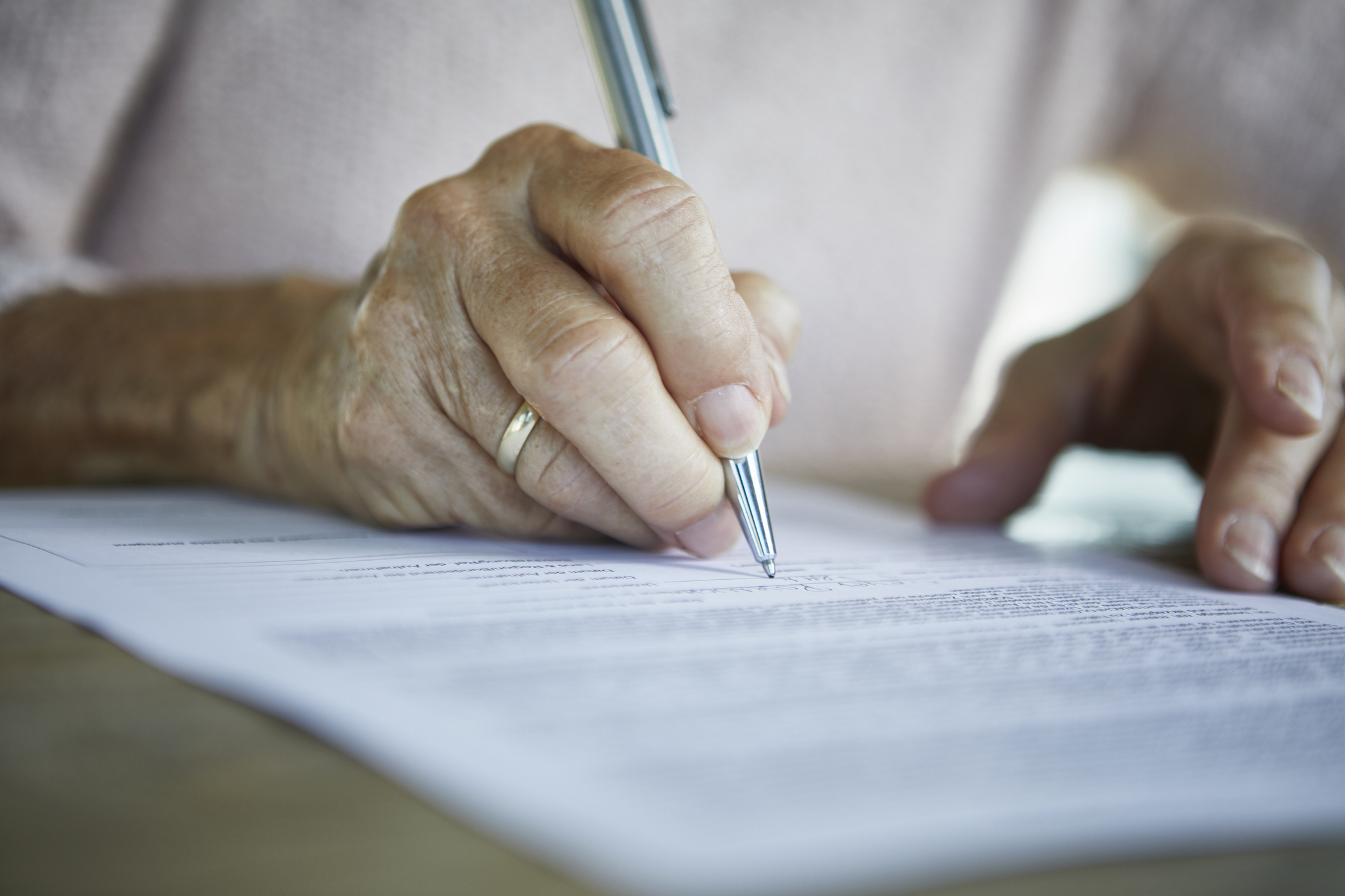 A close-up of an elderly person&#x27;s hand holding a pen while signing legal documents. The person&#x27;s hand displays a gold ring