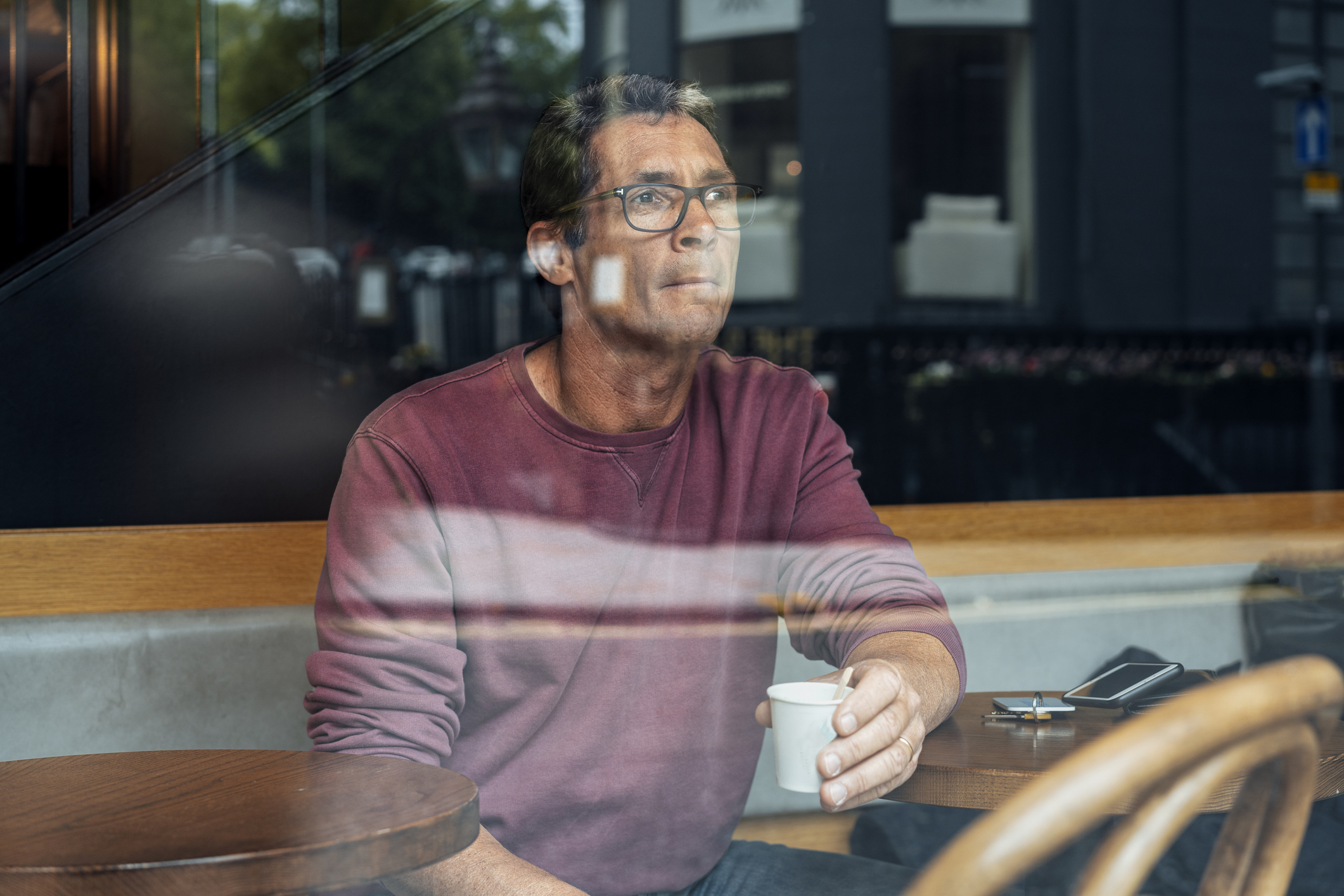 Man wearing glasses and a sweatshirt sitting in a cafe, holding a cup of coffee and looking out the window