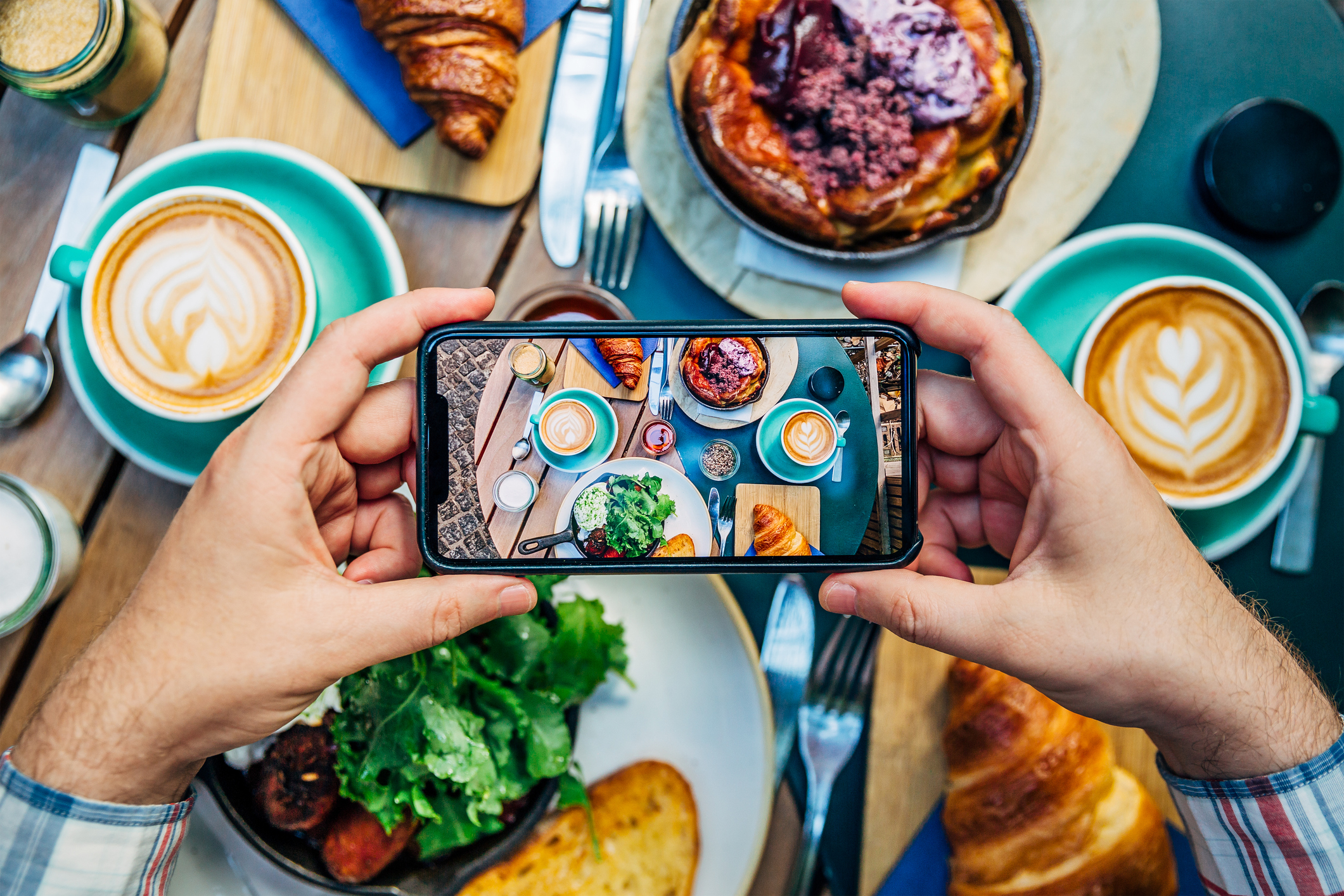 Person photographing a table with croissants, coffee, and various dishes