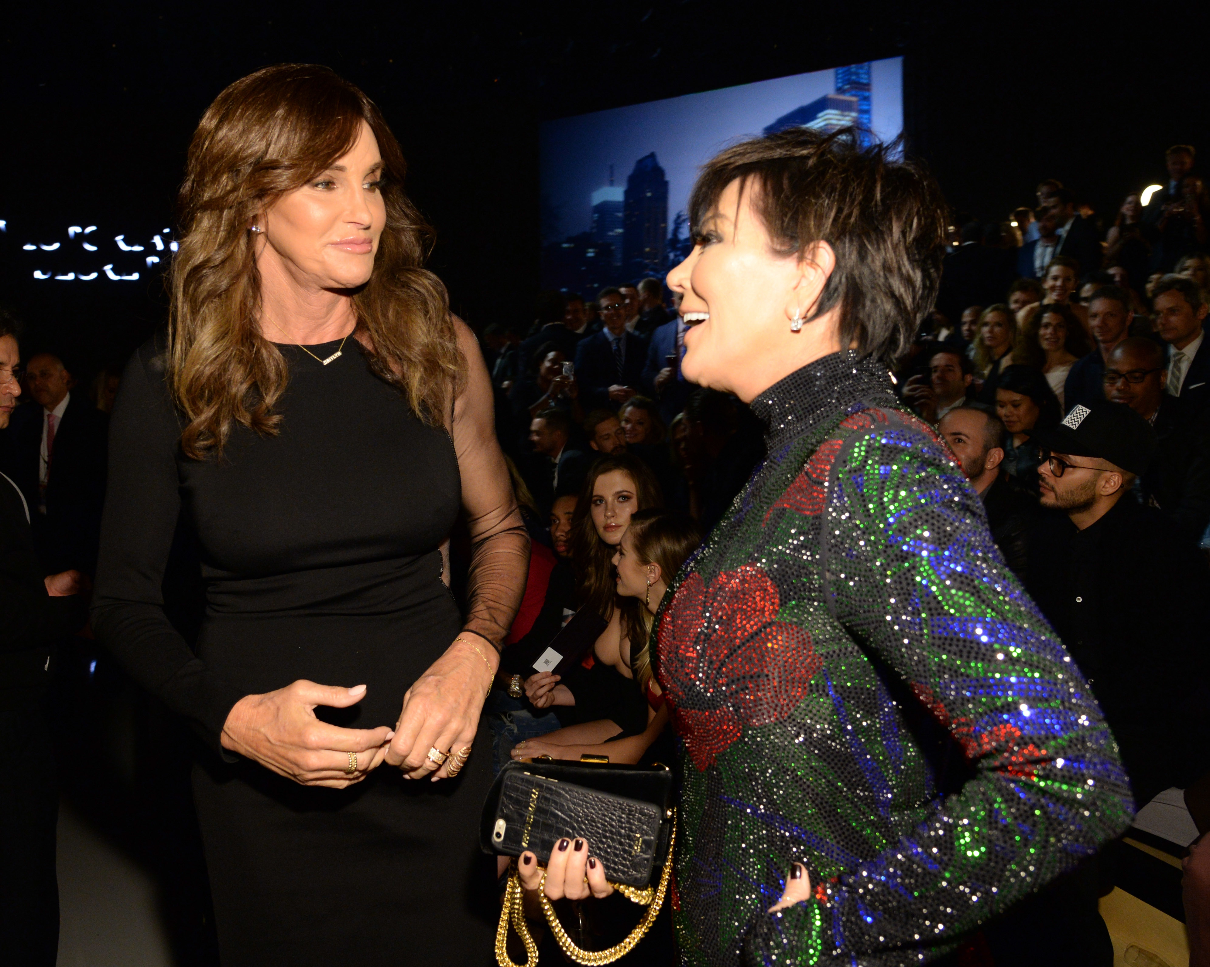 Caitlyn Jenner and Kris Jenner are talking at a public event, with Caitlyn in a black dress and Kris in a sparkling, multi-colored dress