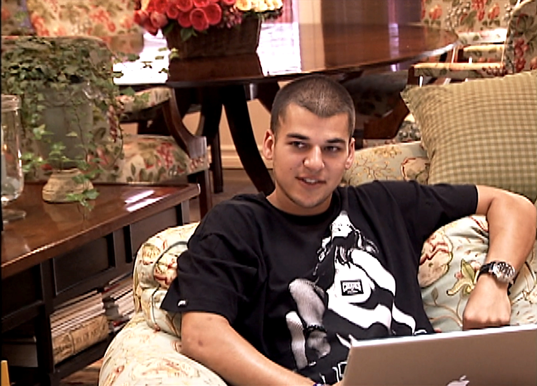 Rob Kardashian sitting on an armchair with a laptop, wearing a graphic t-shirt, in a floral-decorated living room