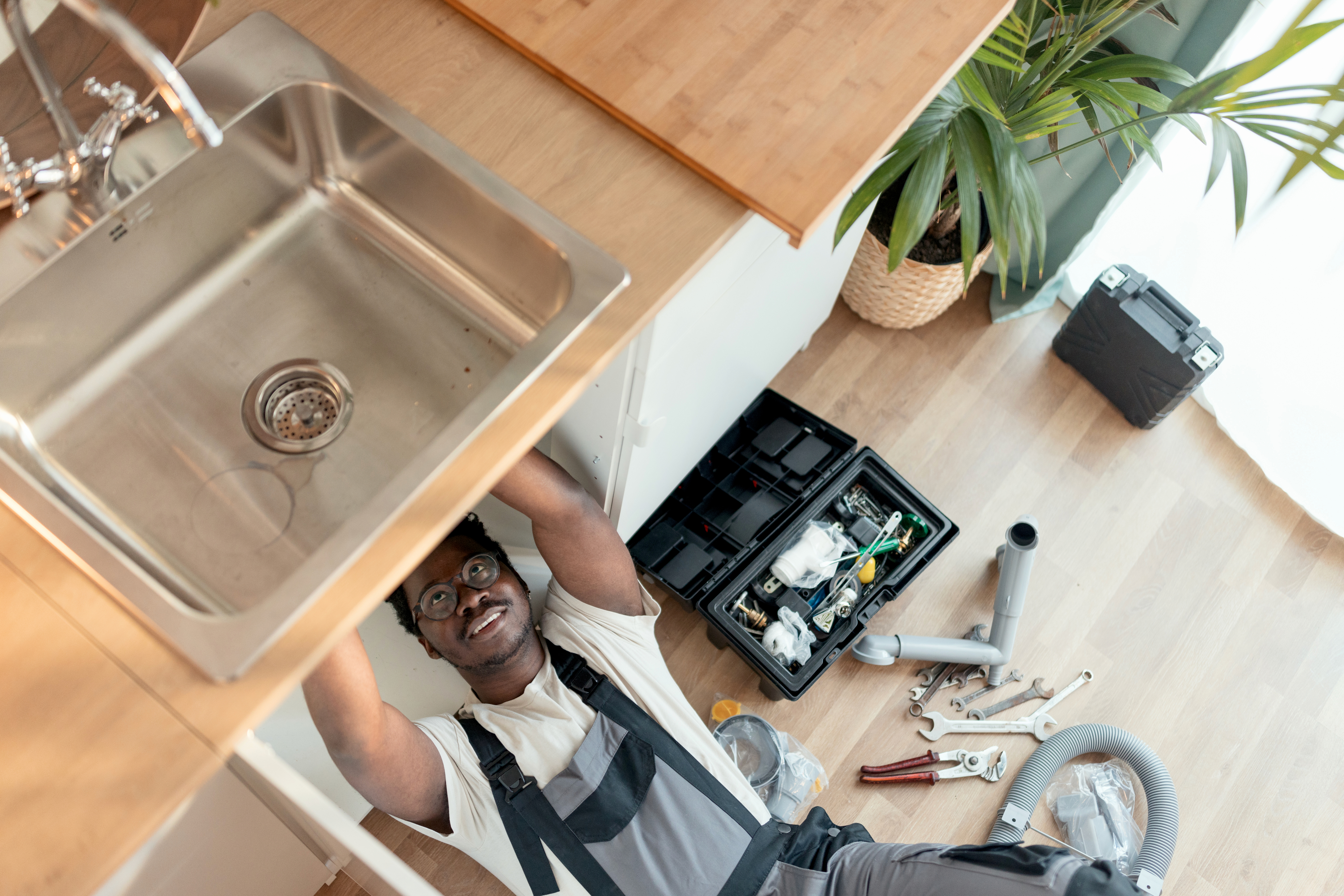 A plumber is lying on his back fixing a sink. Tools and equipment are spread out on the floor next to him, with a plant and a toolbox nearby