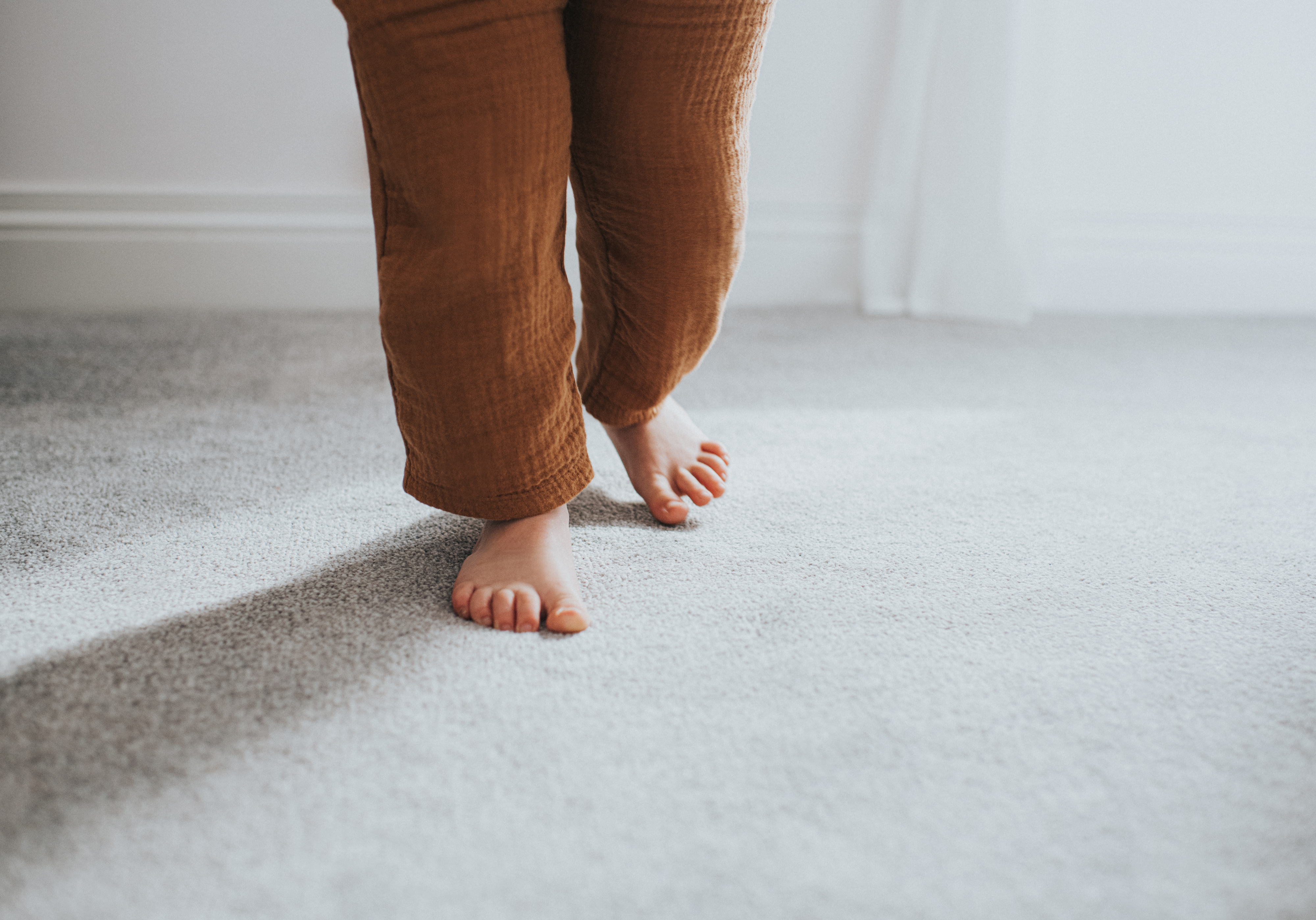 Close-up of a child&#x27;s feet walking on a soft, gray carpet indoors. The child&#x27;s legs are partially visible, wearing loose-fitting pants