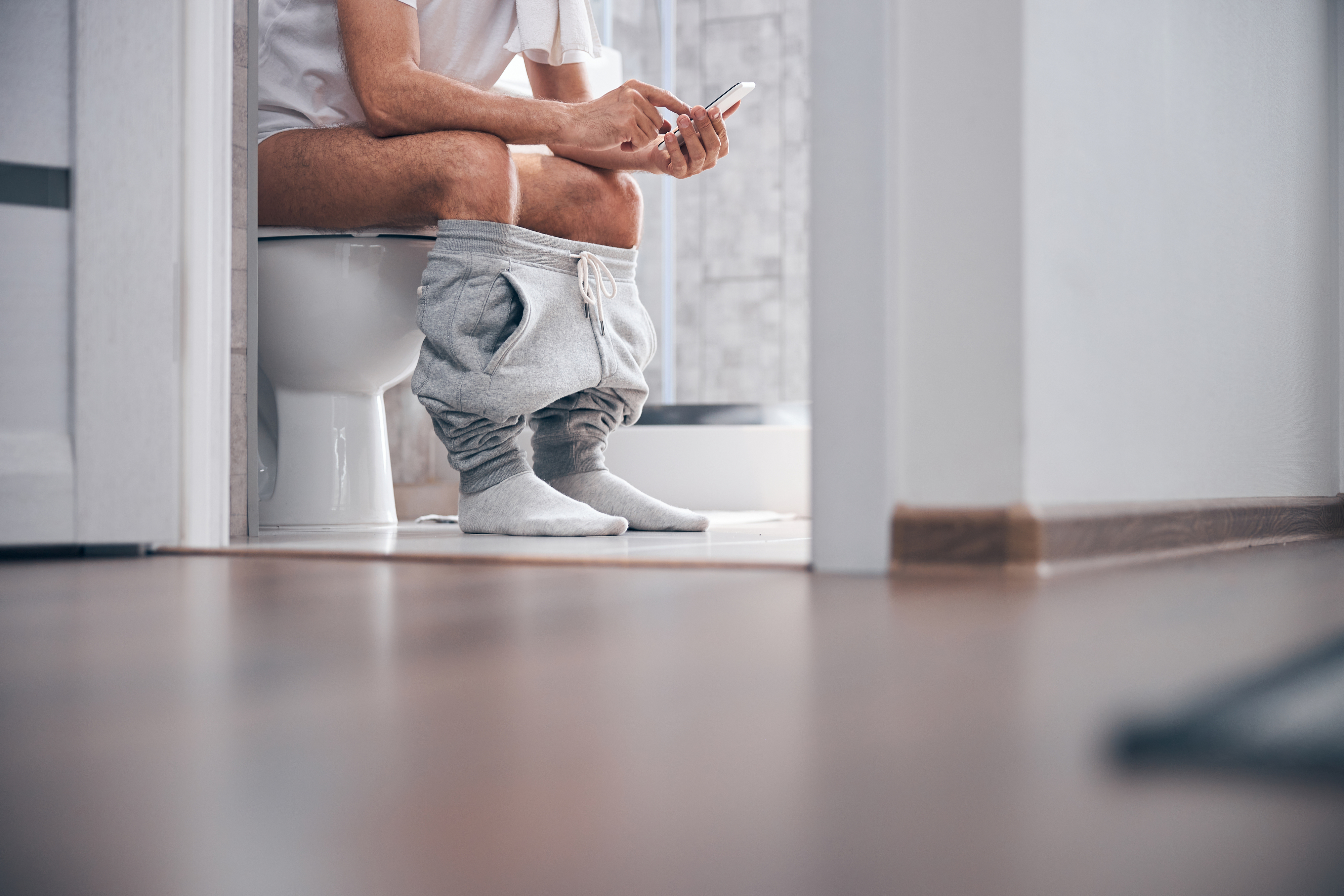 A person sits on a toilet with pants down, looking at a smartphone. The setting appears to be a bathroom with a modern design