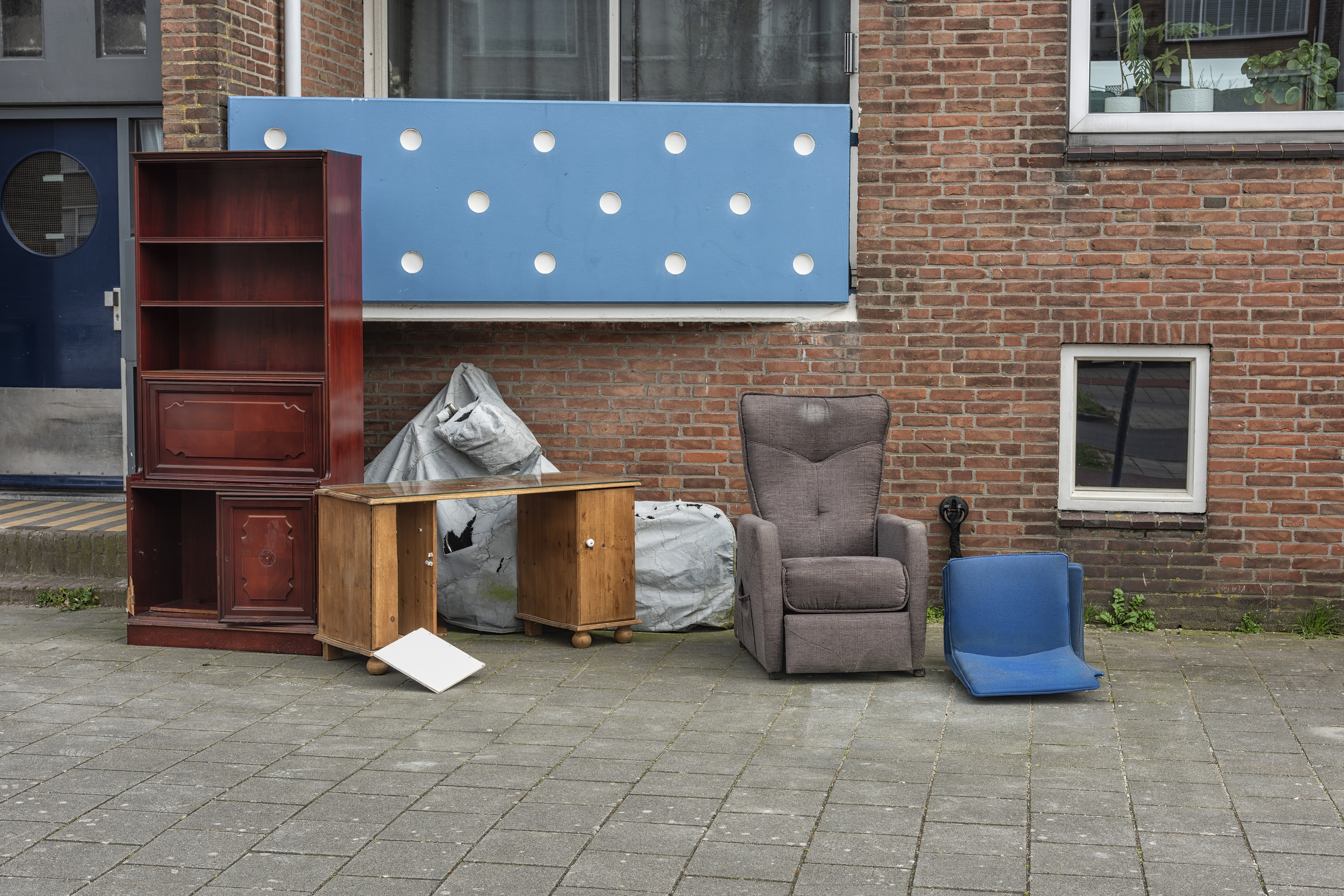 Various pieces of old, discarded furniture, including a bookshelf, cabinets, an armchair, and a blue seat, placed outside a brick building on the sidewalk