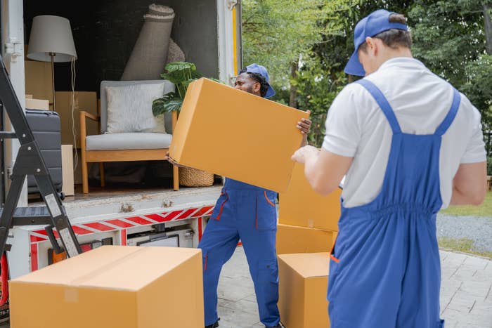 Two movers in blue uniforms load cardboard boxes onto a truck. The truck has furniture inside