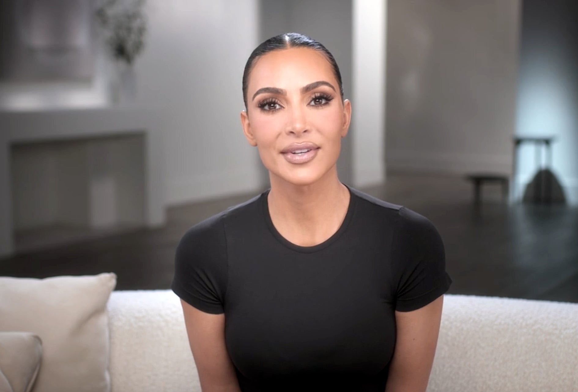 Kim Kardashian sits on a couch wearing a fitted top with her hair slicked back