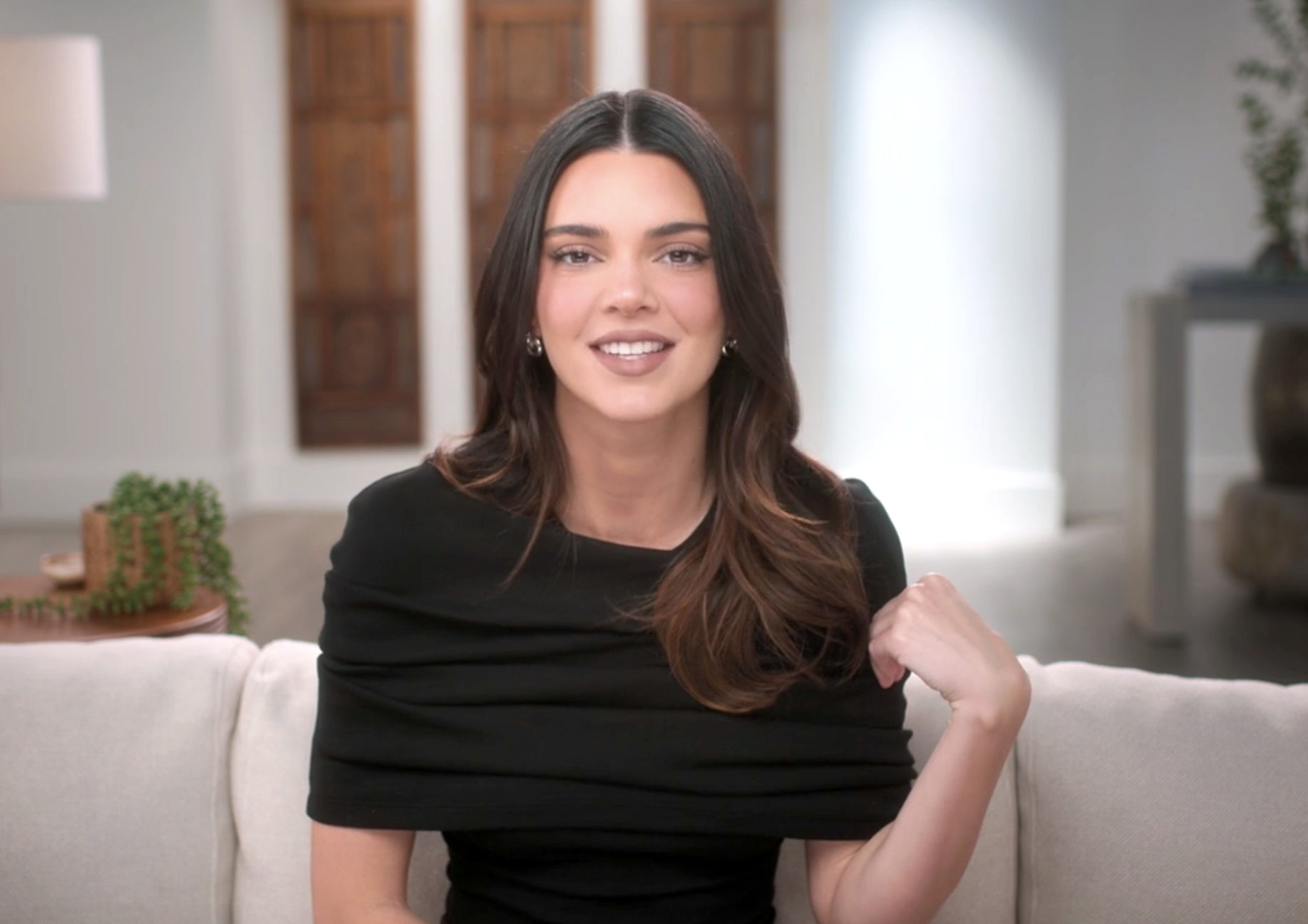 Kendall Jenner sitting on a couch, smiling and wearing a stylish top