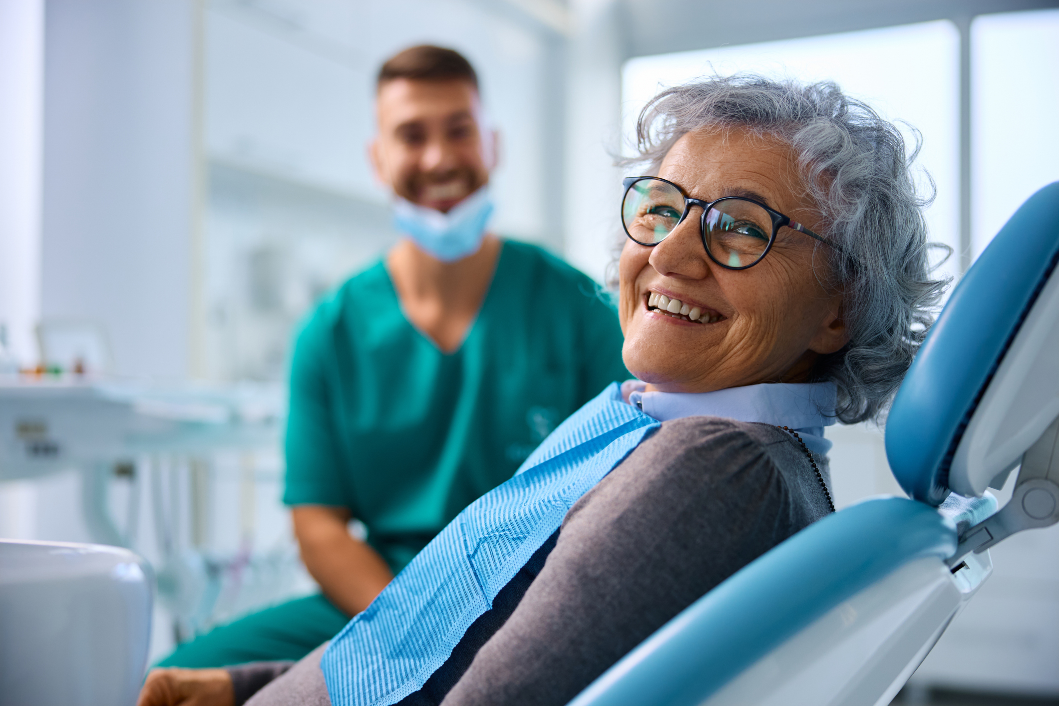 An older woman with glasses smiles sitting in a dentist&#x27;s chair, while a masked dentist in scrubs smiles in the background