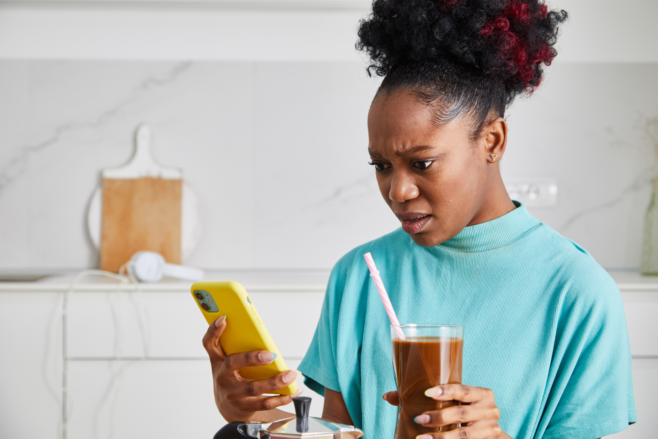 Woman in a turquoise top holds a yellow phone and a glass of iced coffee, looking at the phone with a concerned expression