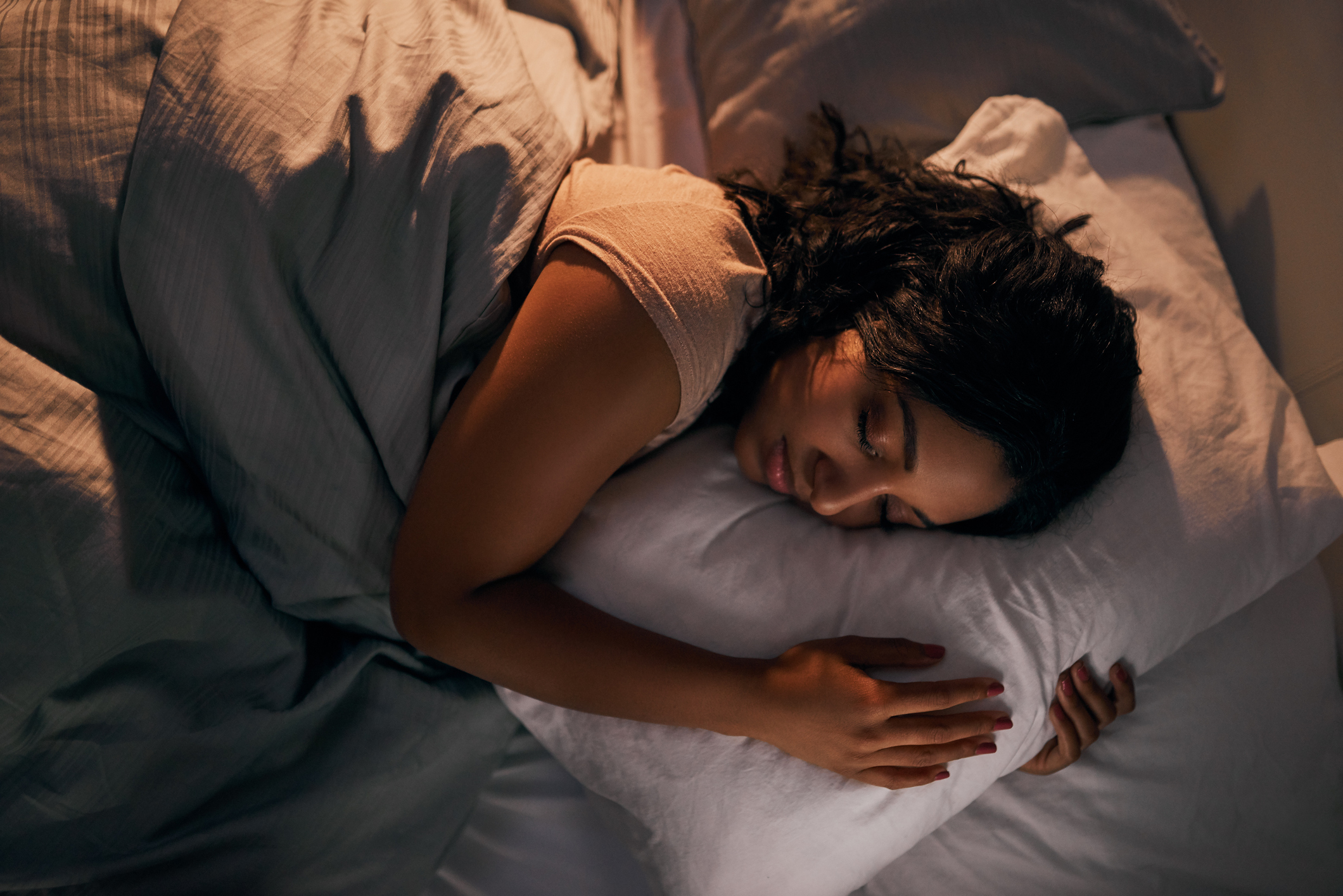 A person with dark hair sleeps peacefully on a bed, hugging a pillow and covered with a blanket