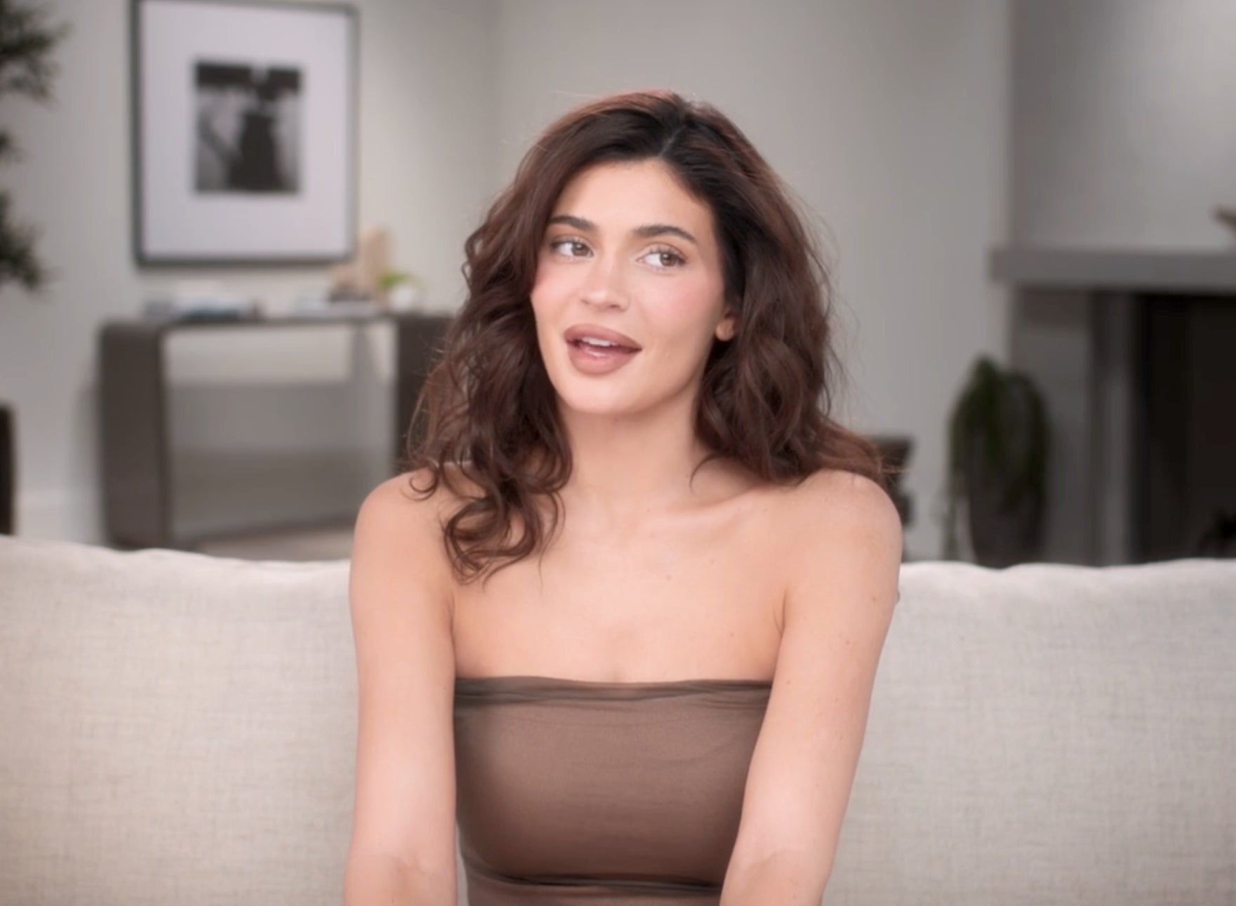 Kylie Jenner sits on a couch wearing a strapless top and speaking during a confessional