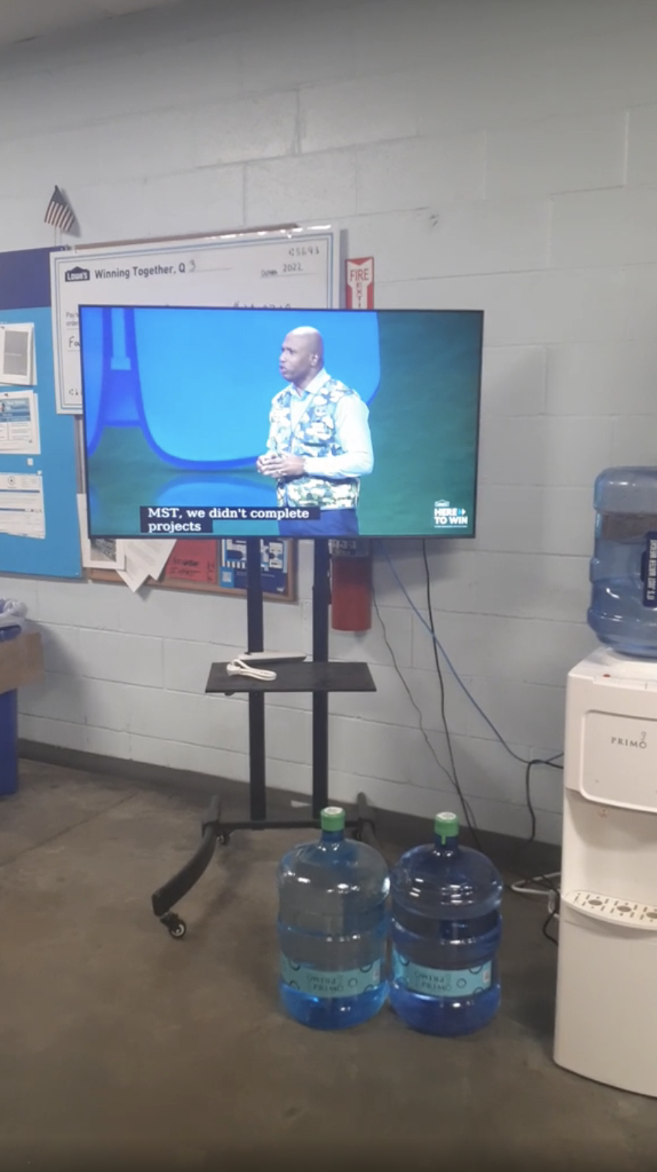 A person is speaking on a TV screen in a room with water coolers and office equipment. The screen displays subtitles but the person&#x27;s name is not visible