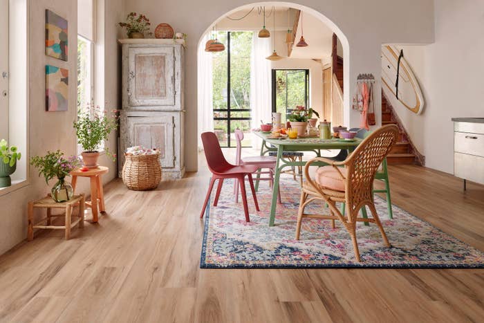 Modern dining area with wooden floors, a mix of colorful chairs around a table, and a bright rug. Large arched doorway opens to greenery. Decor is eclectic