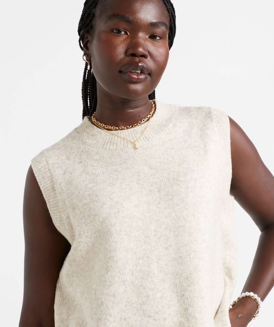 Woman in sleeveless knit top with braided hair, wearing hoop earrings, a necklace, and a bracelet, modeling for a clothing brand