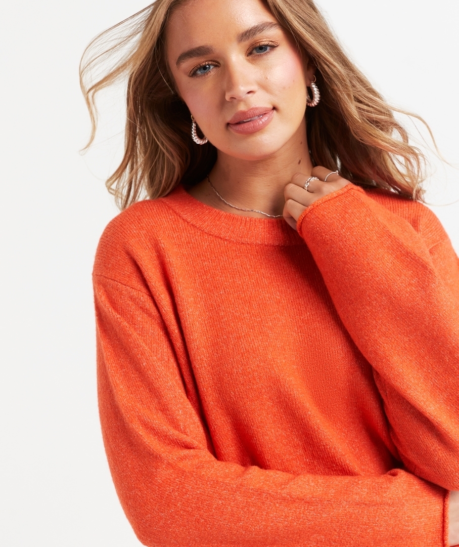 A woman with wavy hair, wearing a long-sleeve, knitted sweater poses with a relaxed expression. She accessorizes with hoop earrings and a delicate necklace