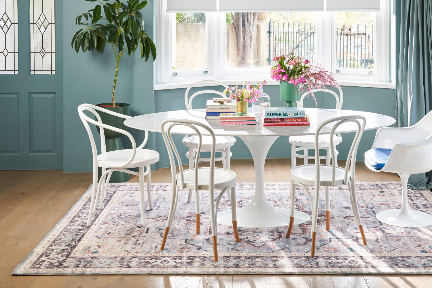 A modern dining room setup featuring a round white table, white chairs, a vase with flowers, and a stack of books. The room has a large window and a decorative rug