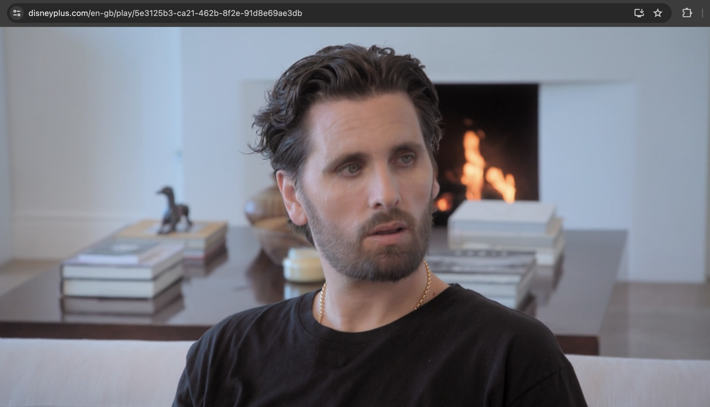 Scott Disick sits indoors with a pensive expression, wearing a casual black T-shirt. A fireplace and stacked books are visible in the background