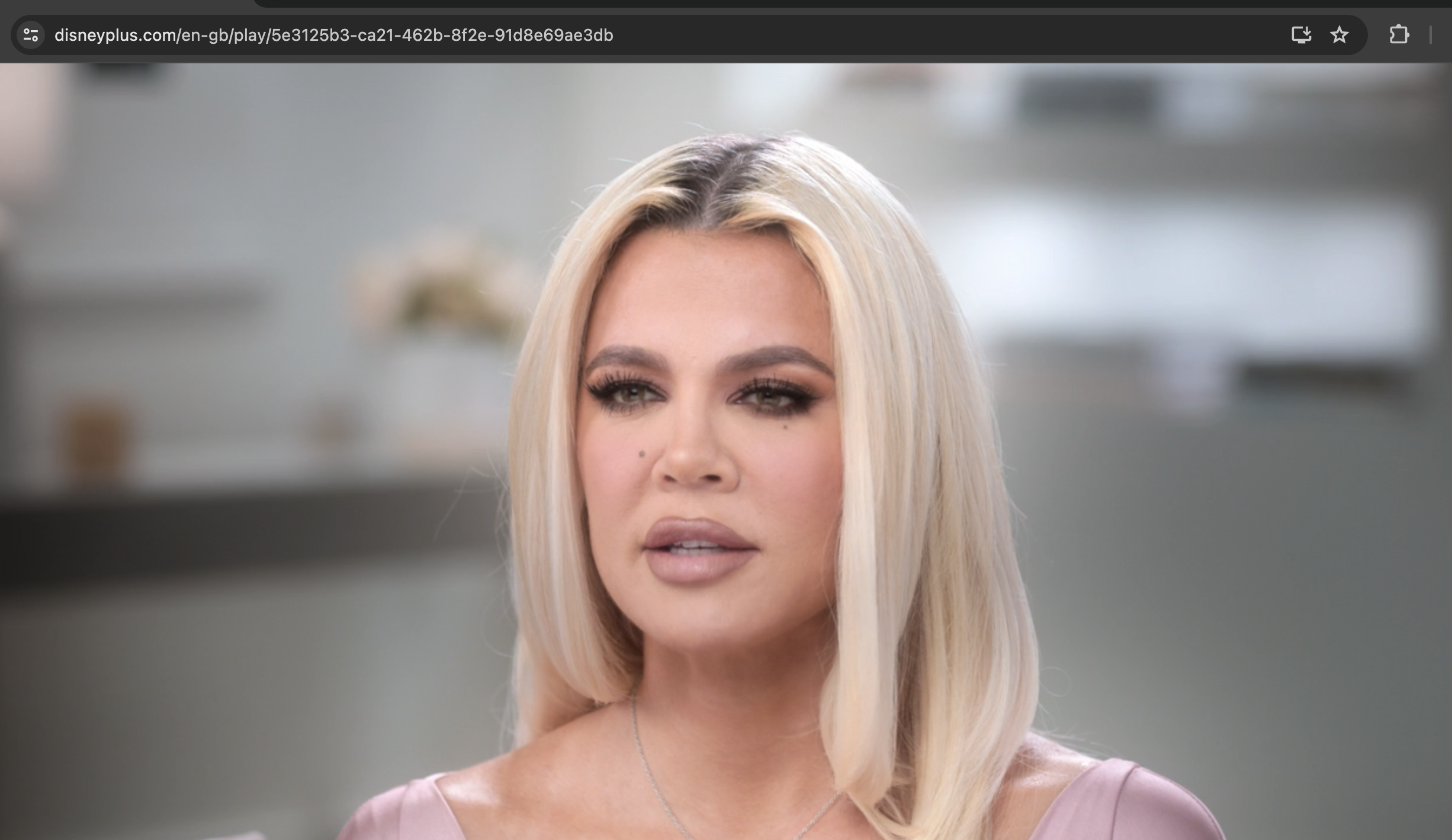 Khloé Kardashian in a close-up shot from an interview, wearing a light-colored top and a heart pendant necklace