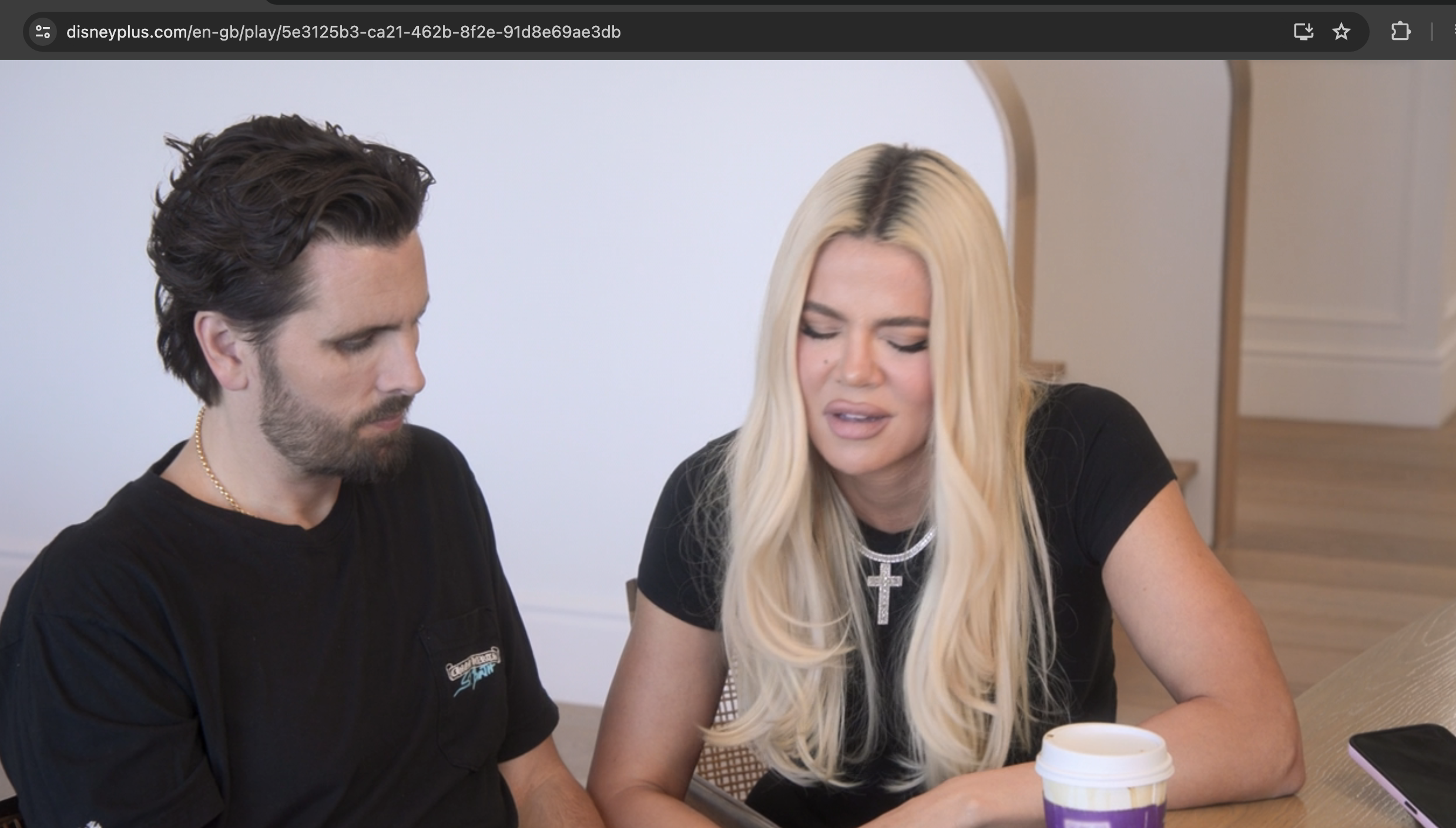 Scott Disick and Khloe Kardashian are seated indoors, looking at a smartphone. Khloe has straight blonde hair and a chunky cross necklace; a coffee cup is beside her