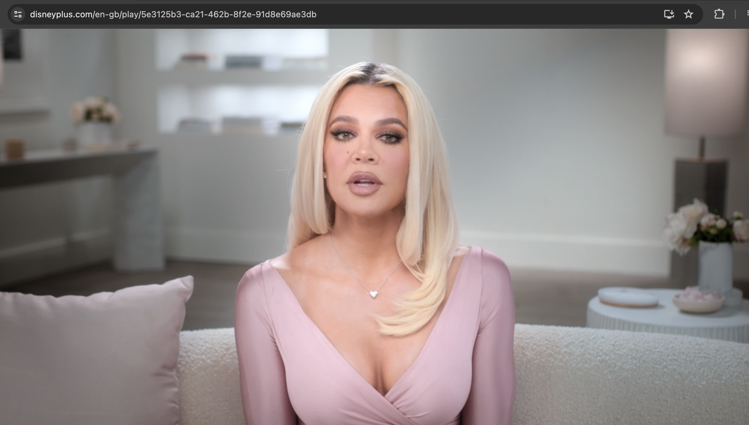 Khloé Kardashian sits on a couch in a stylish light pink dress, speaking directly to the camera in a scene from &quot;The Kardashians&quot; on Disney+