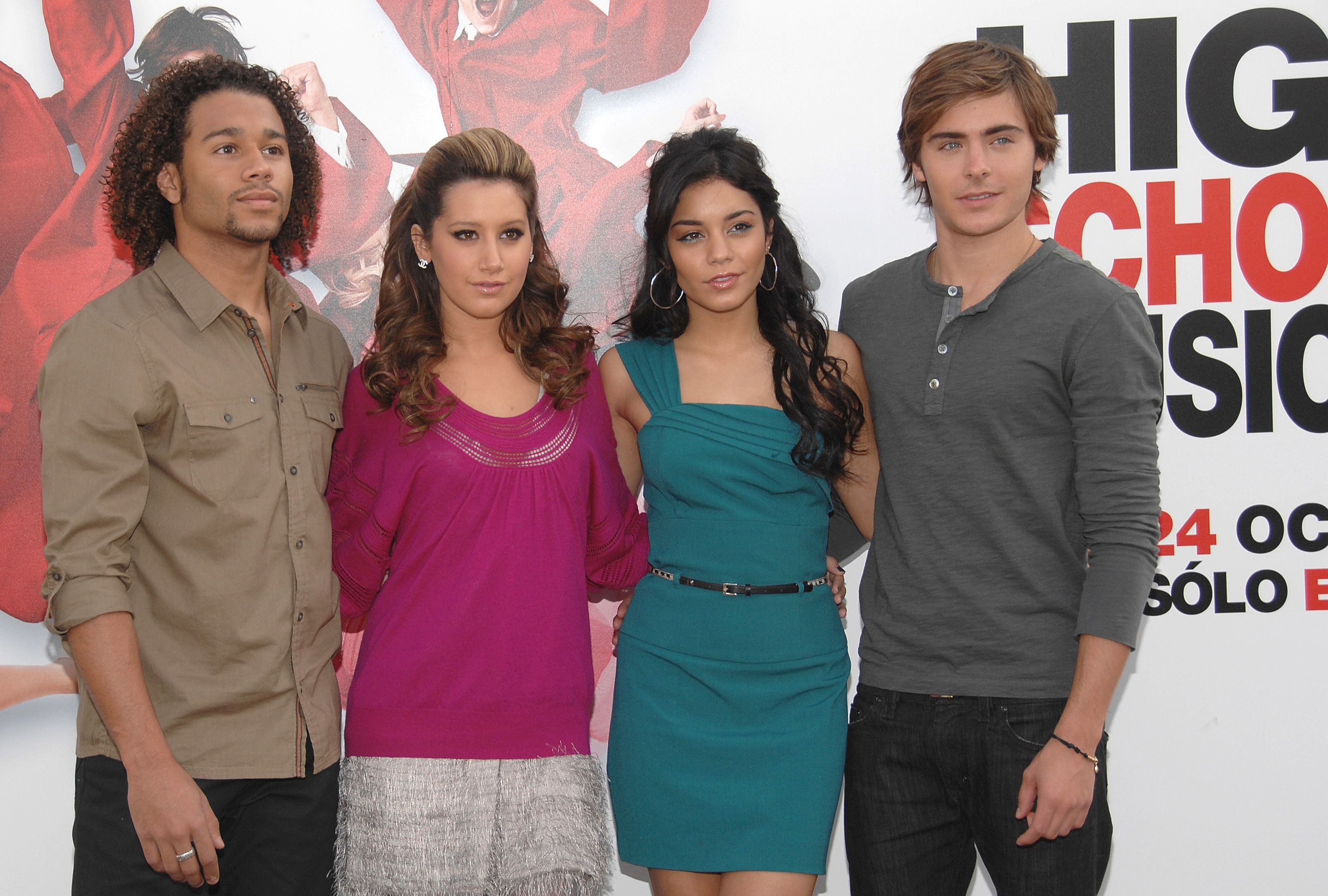 Corbin Bleu, Ashley Tisdale, Vanessa Hudgens, and Zac Efron stand together in casual outfits in front of a &quot;High School Musical&quot; poster
