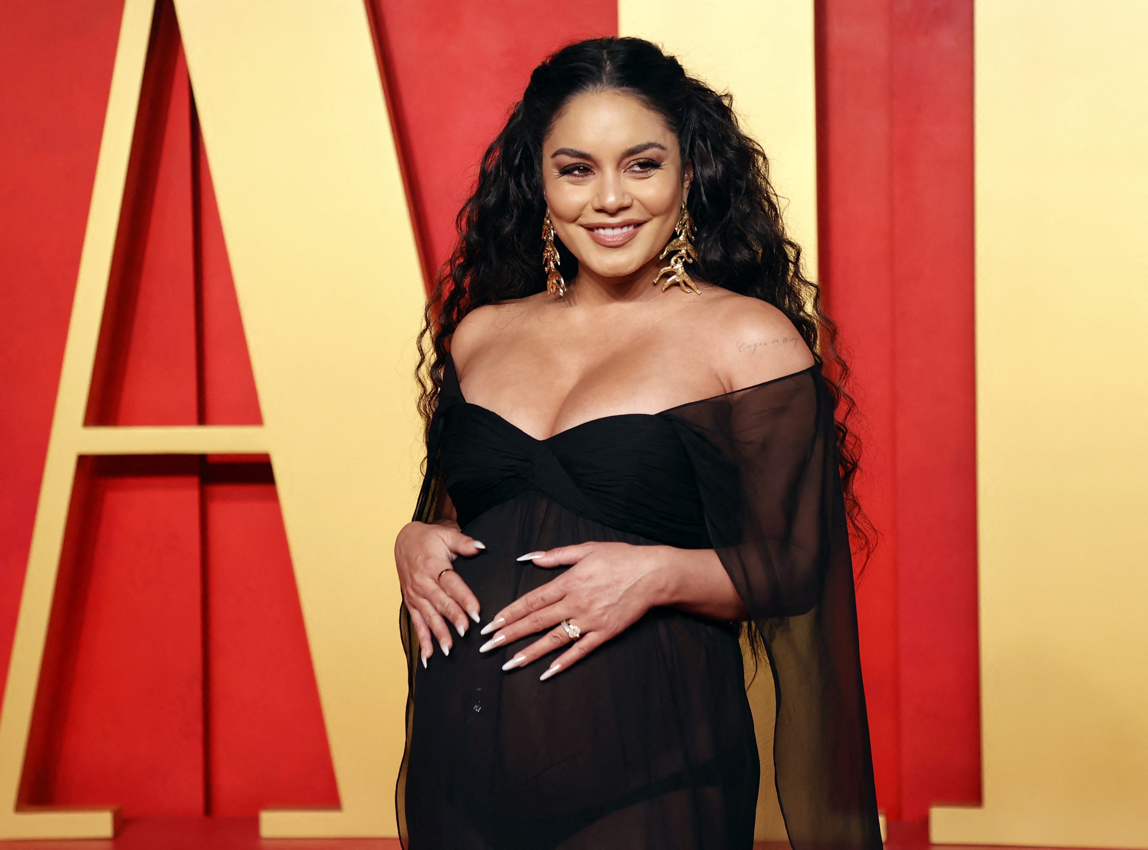 Vanessa Hudgens poses on the red carpet in a sheer dress, holding her baby bump and smiling