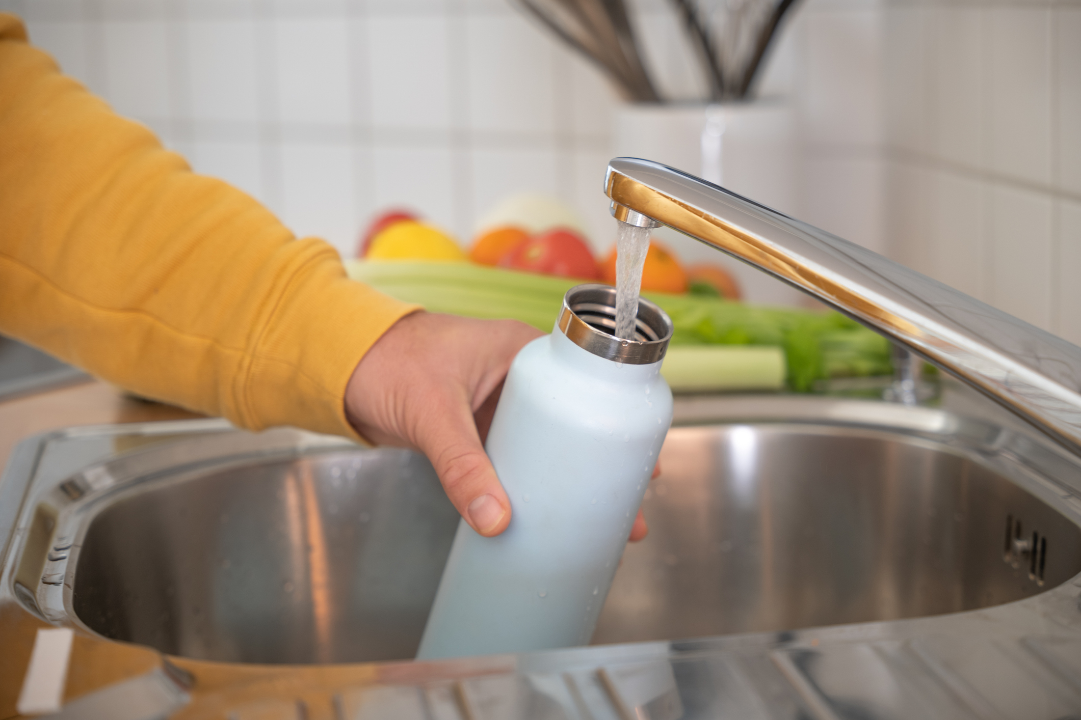A person in a yellow sleeve fills a reusable water bottle from a kitchen faucet. Fruits and vegetables are in the background