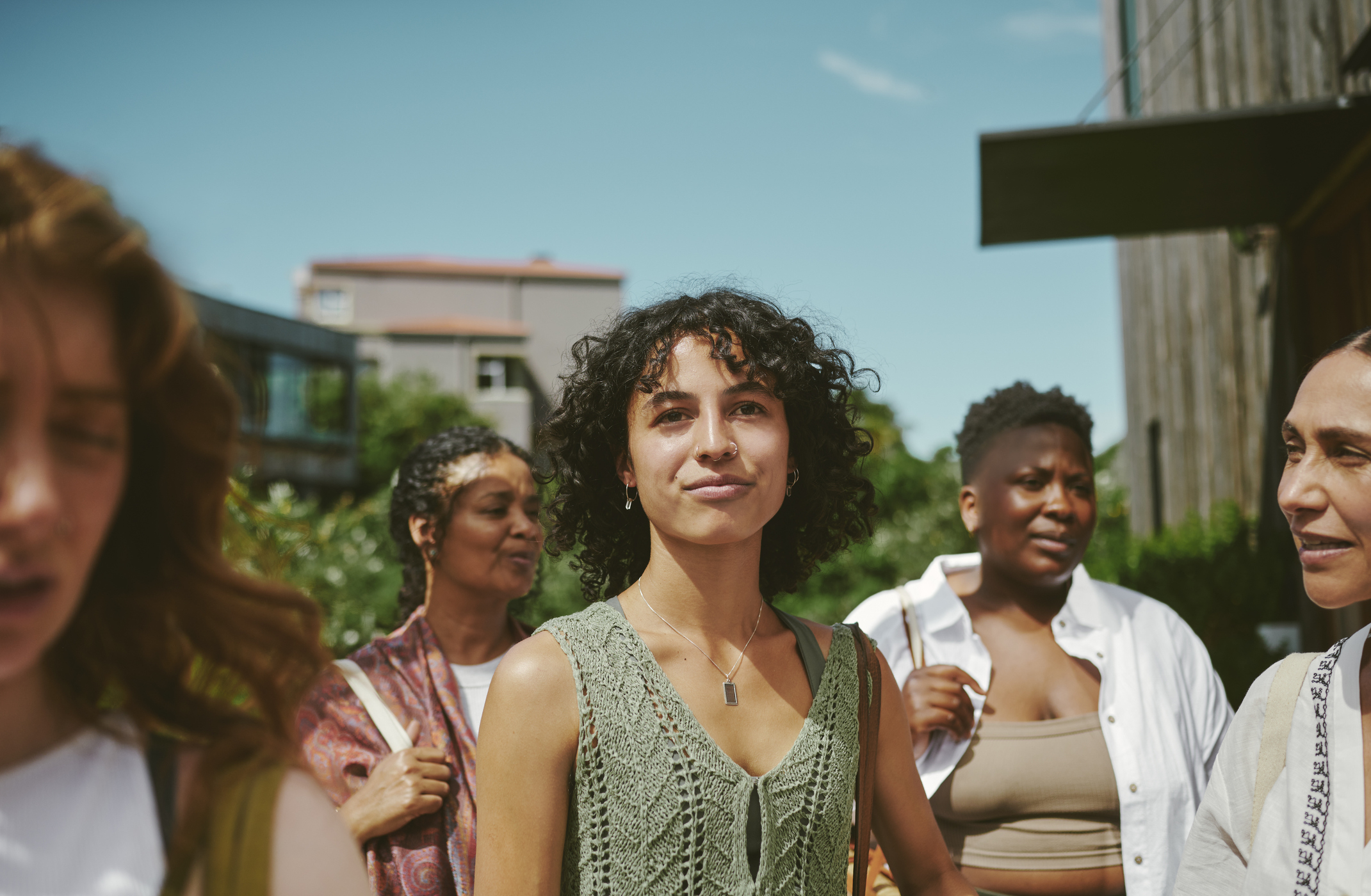 Five people are standing outdoors with buildings and greenery in the background. A person in a green top is at the center of the image