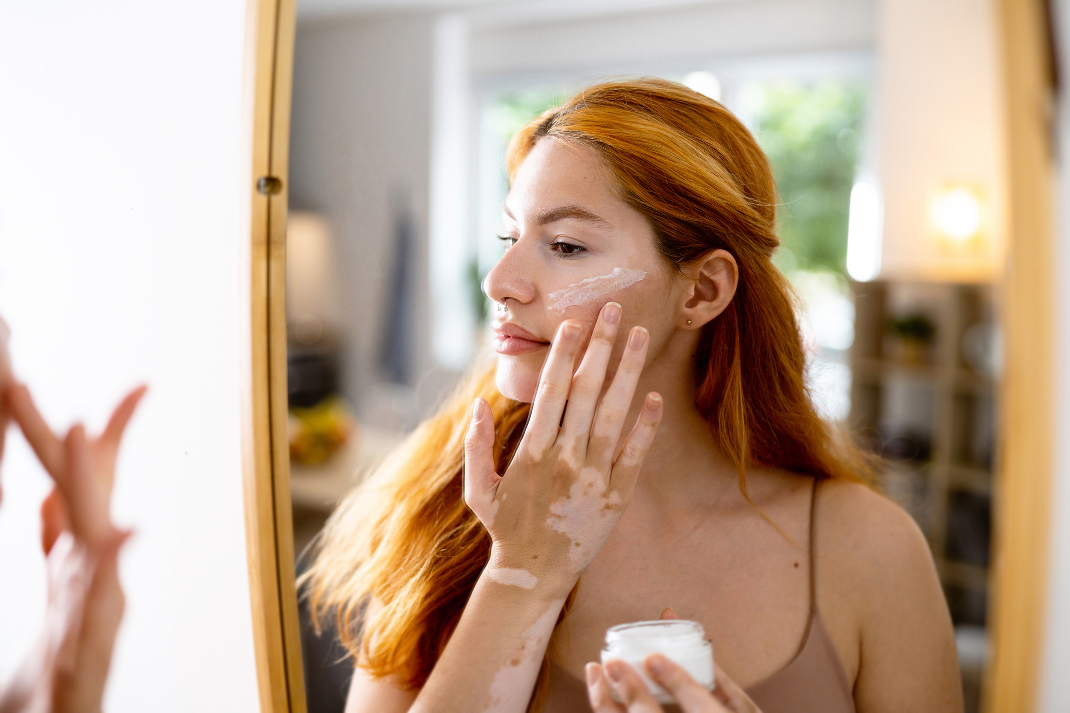 A woman is applying face cream while looking in the mirror. She has long, straight hair and is wearing a sleeveless top. No names are provided