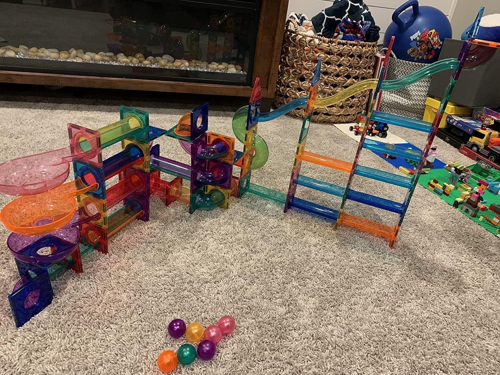 Colorful marble run set with tracks and structures set up on a carpeted floor. Various toys and a basket of toys are visible in the background