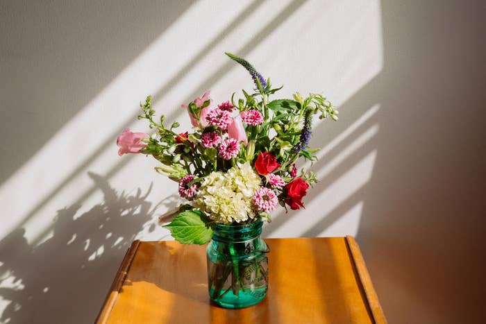A small bouquet of various flowers in a green jar sits on a wooden table in sunlight