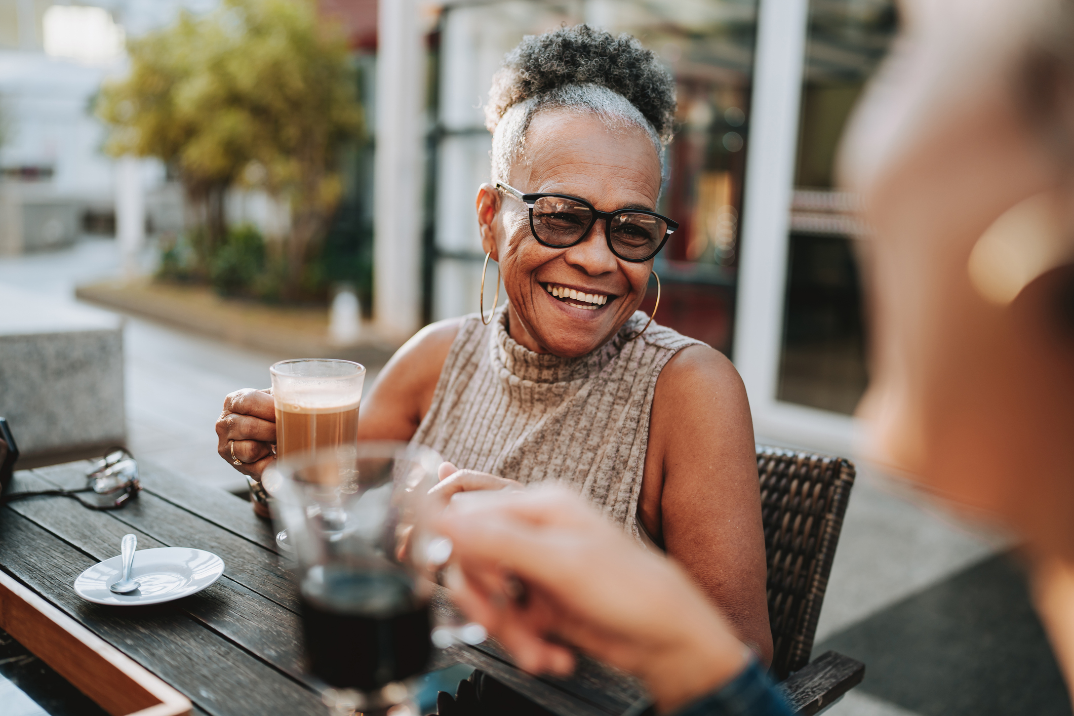 An older woman with glasses and a sleeveless top smiles while holding a cup of coffee, sitting at an outdoor table with another person