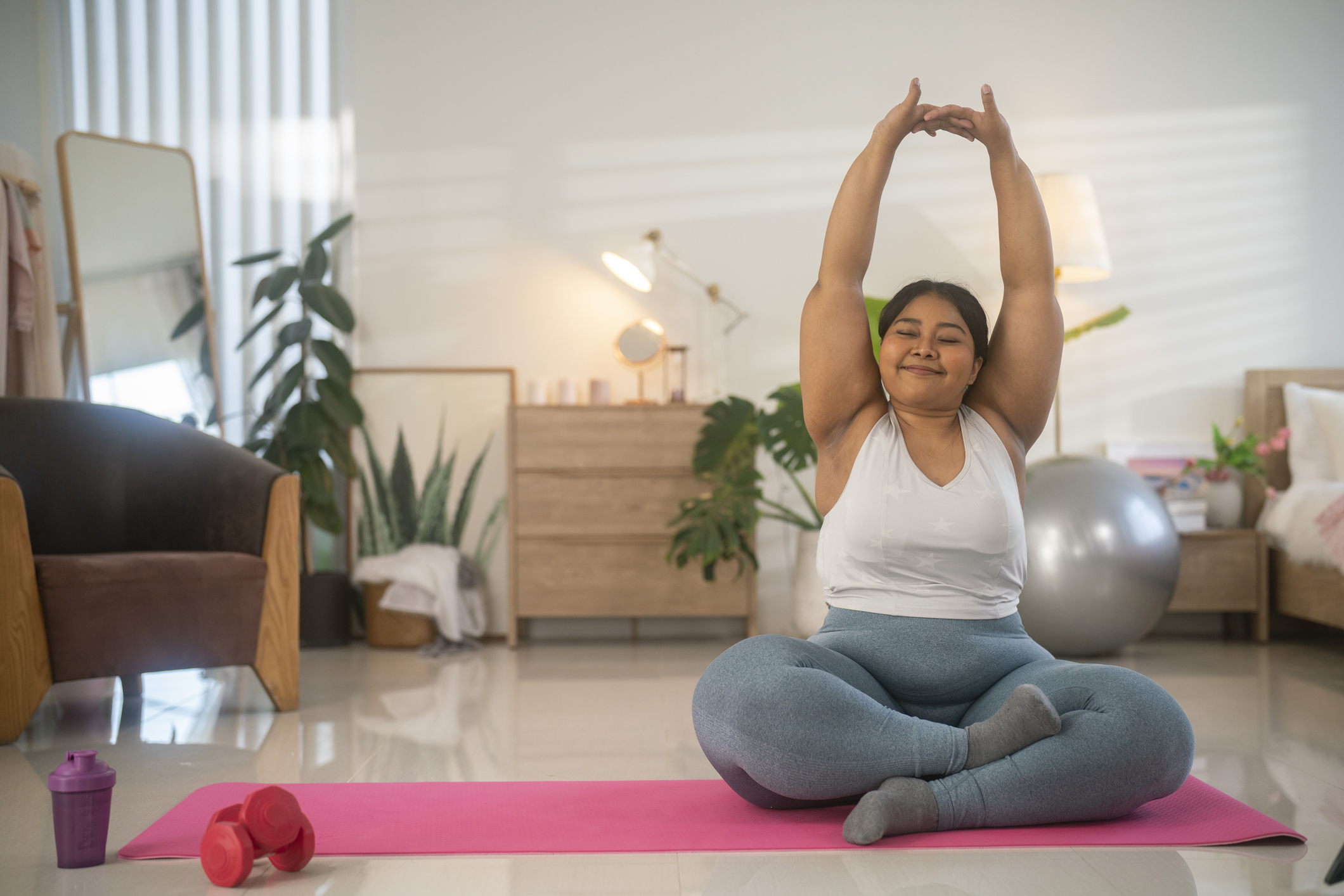 Person doing yoga at home, seated on a pink mat with legs crossed, arms stretched overhead, smiling. A dumbbell, a water bottle, and a large exercise ball are nearby